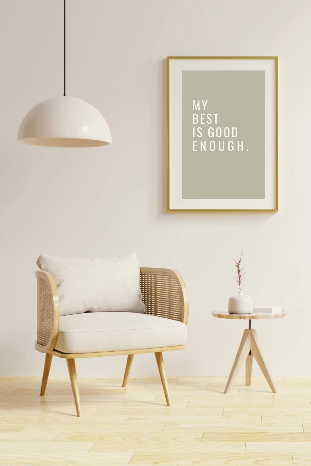 Minimalist living room corner with a white armchair, a small round wooden side table with a white vase and a branch, and a framed quote on the wall that says 'My best is good enough.' A white hanging pendant light is above the armchair.