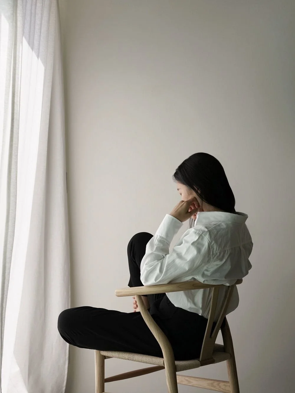 A woman sitting on a wooden chair near a window with white curtains, with her head resting on her hand in a contemplative pose.