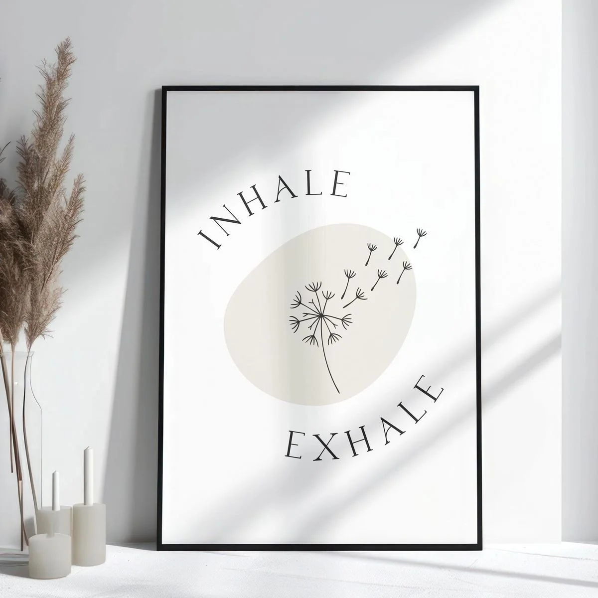 Minimalist framed poster with the words "Inhale" and "Exhale" and a dandelion illustration, placed on a white wall near dried pampas grass and candles.