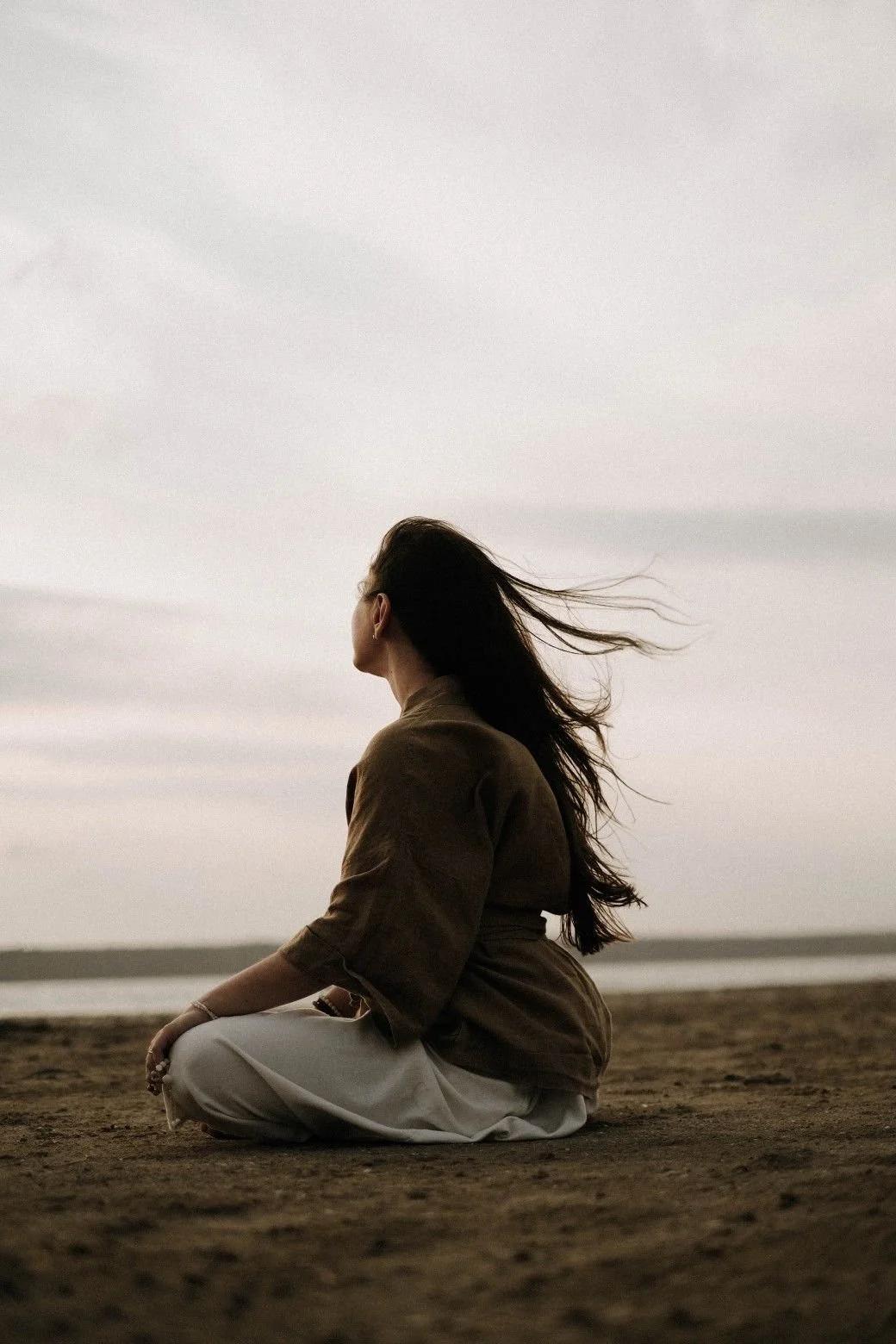 A woman sitting cross-legged on a sandy beach, facing sideways with her hair blowing in the wind, during overcast weather.