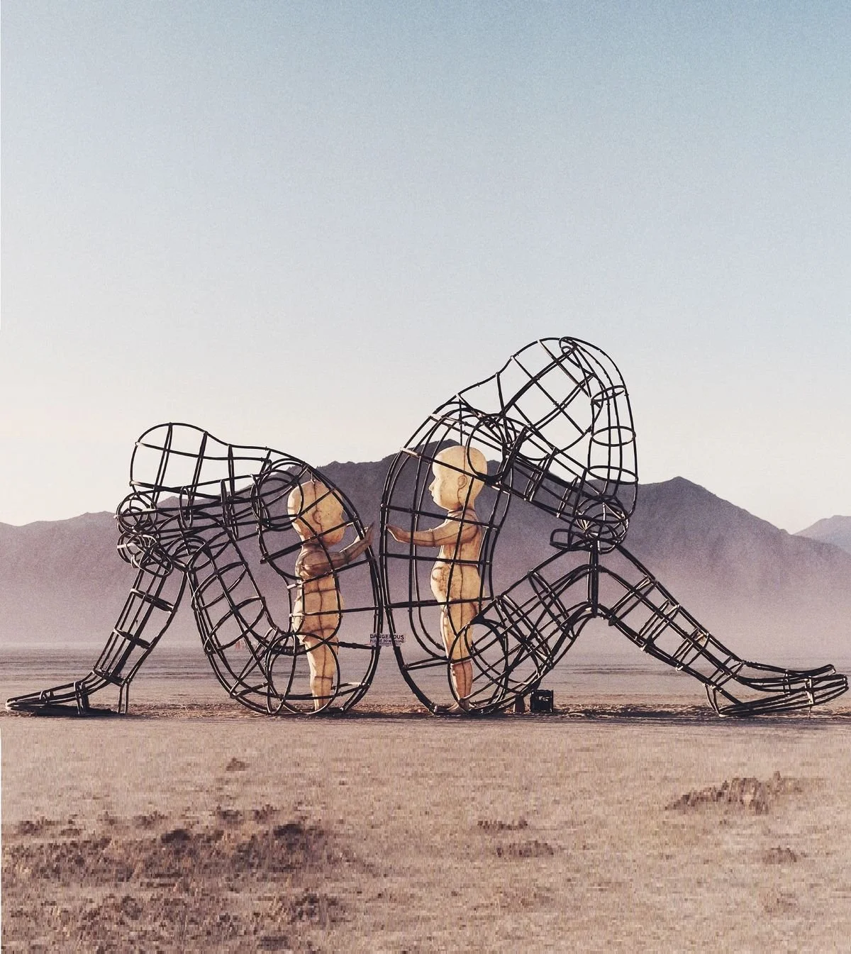Large metal sculpture in a desert landscape showing multiple human figures in different postures, with mountain range in the background.