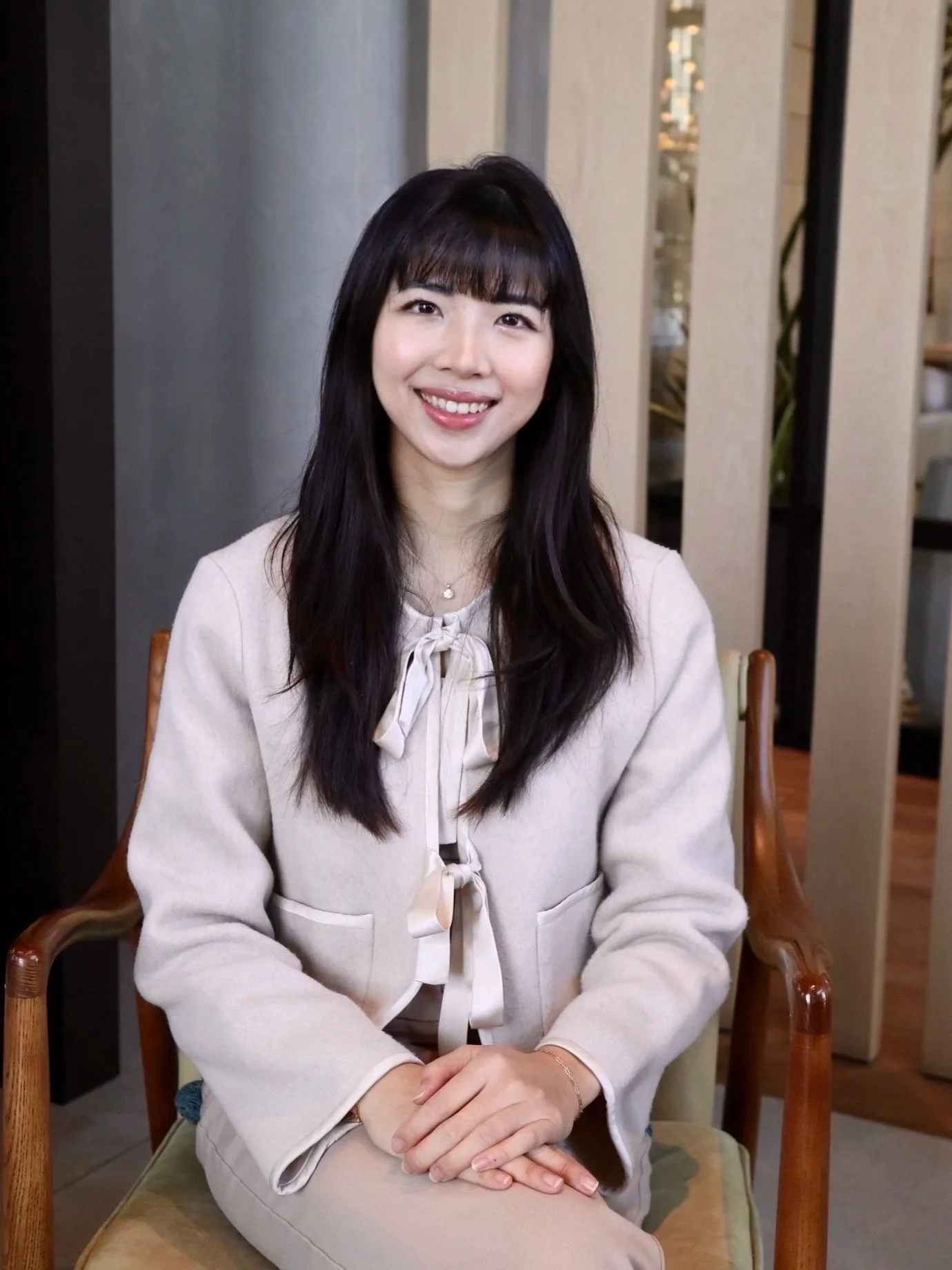 A female anxiety therapist in NYC with long black hair and bangs, smiling and sitting on a wooden chair in an indoor setting with modern decor.