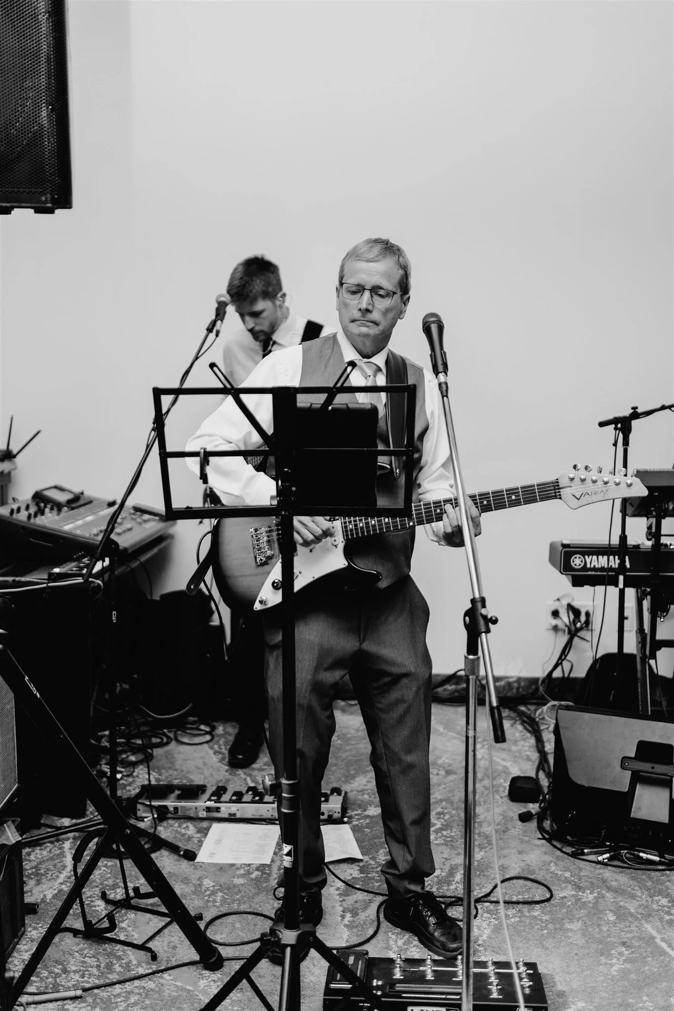 Black and white photo of a man playing an electric guitar and singing into a microphone, with another musician in the background at a music performance.