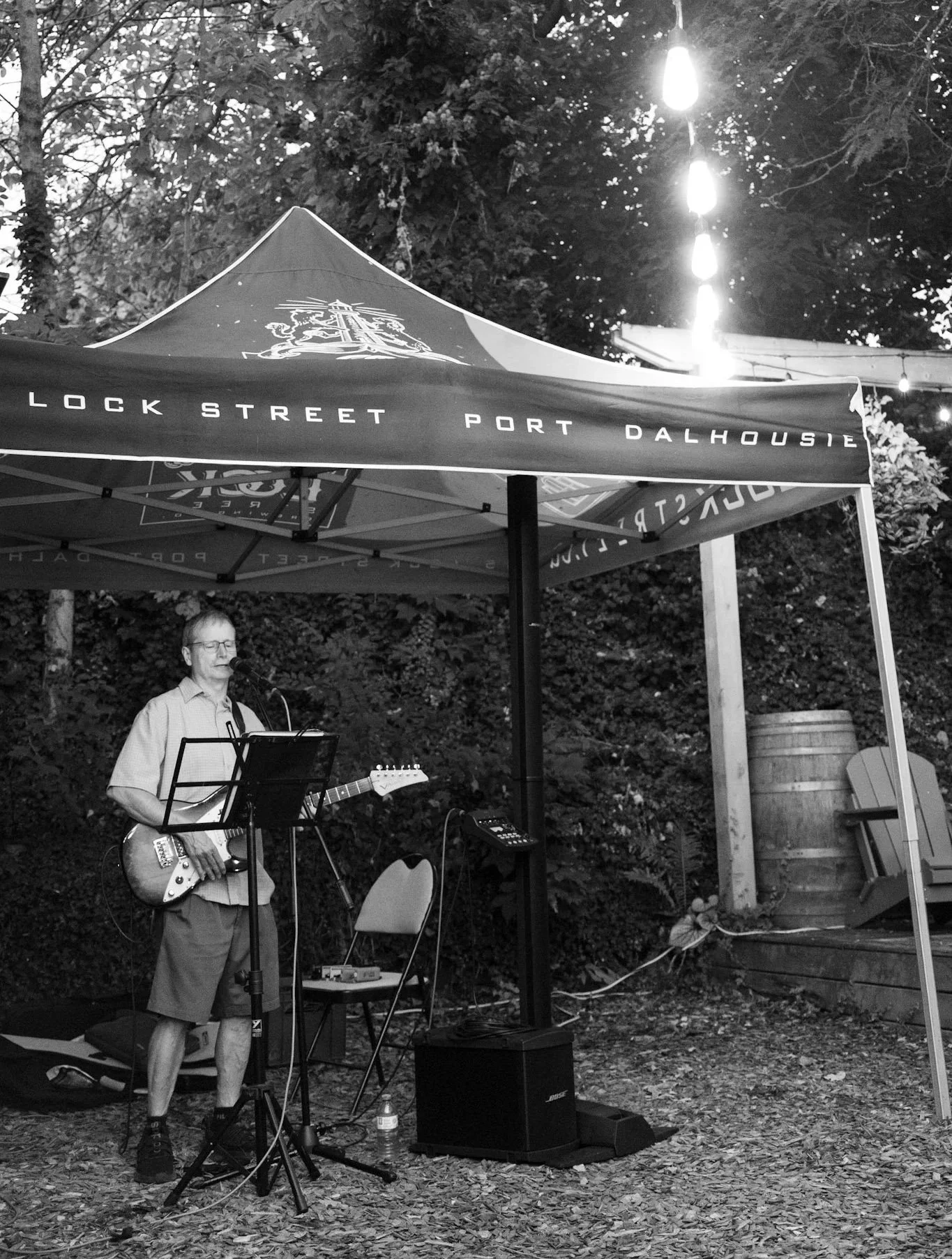 A man playing guitar and singing into a microphone under a canopy with the words "Lock Street Port Dalhousie." There are string lights hanging above and a wooden barrel to the side with outdoor chairs nearby.
