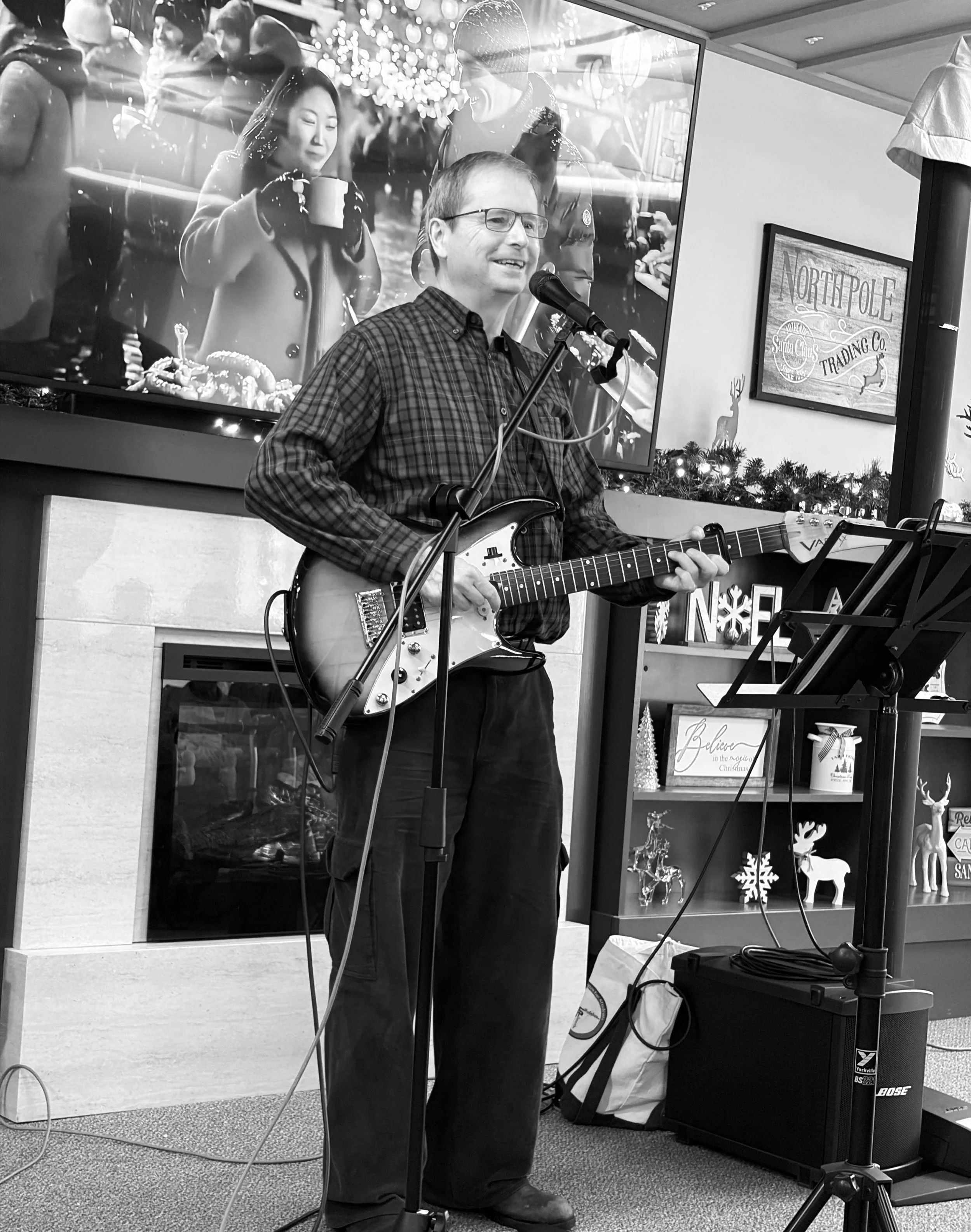 Man wearing glasses playing electric guitar and singing into a microphone at a holiday event, with Christmas decorations and a large television screen in the background.