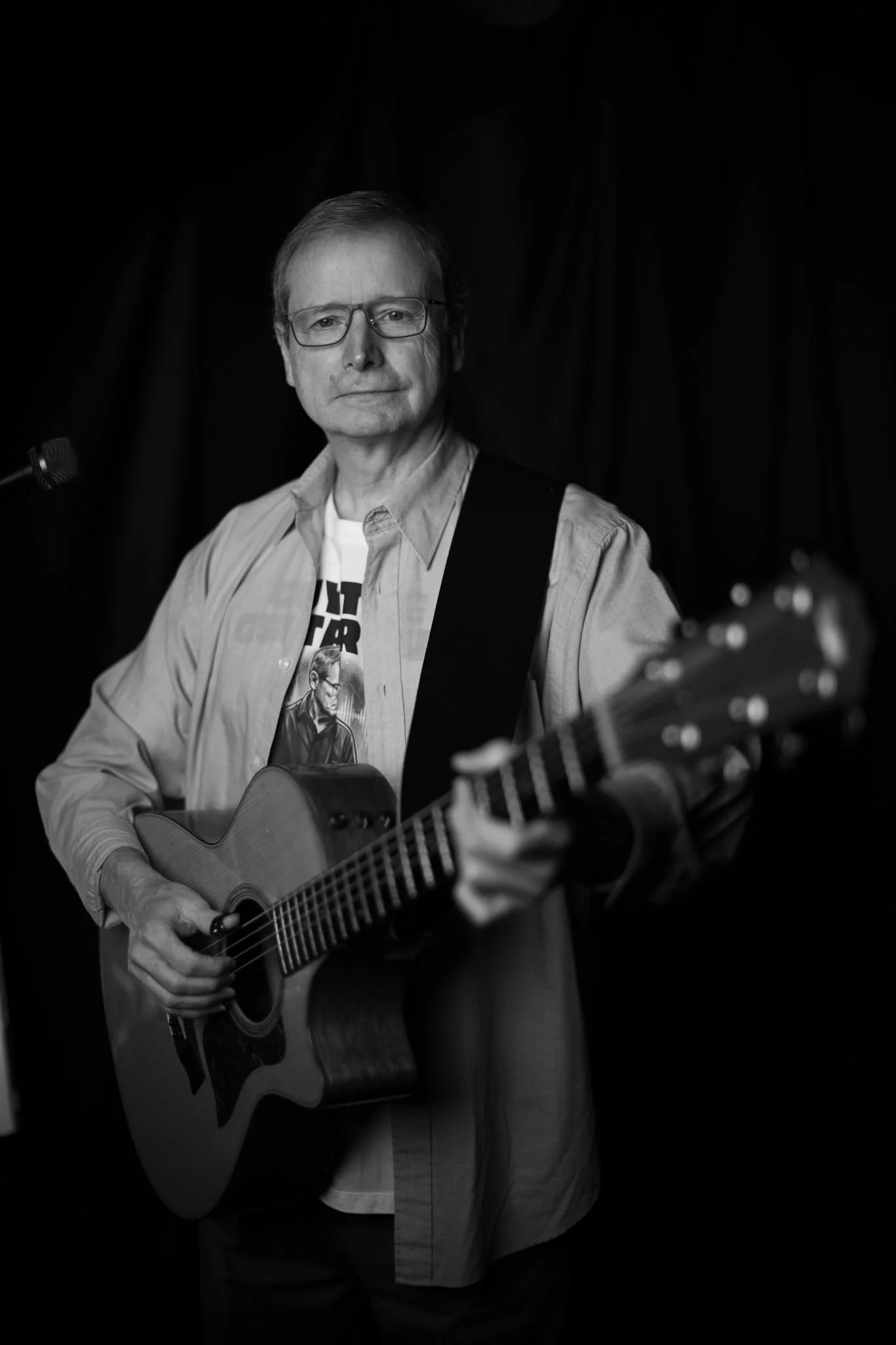 A middle-aged man with glasses holding an acoustic guitar on stage with a dark background.