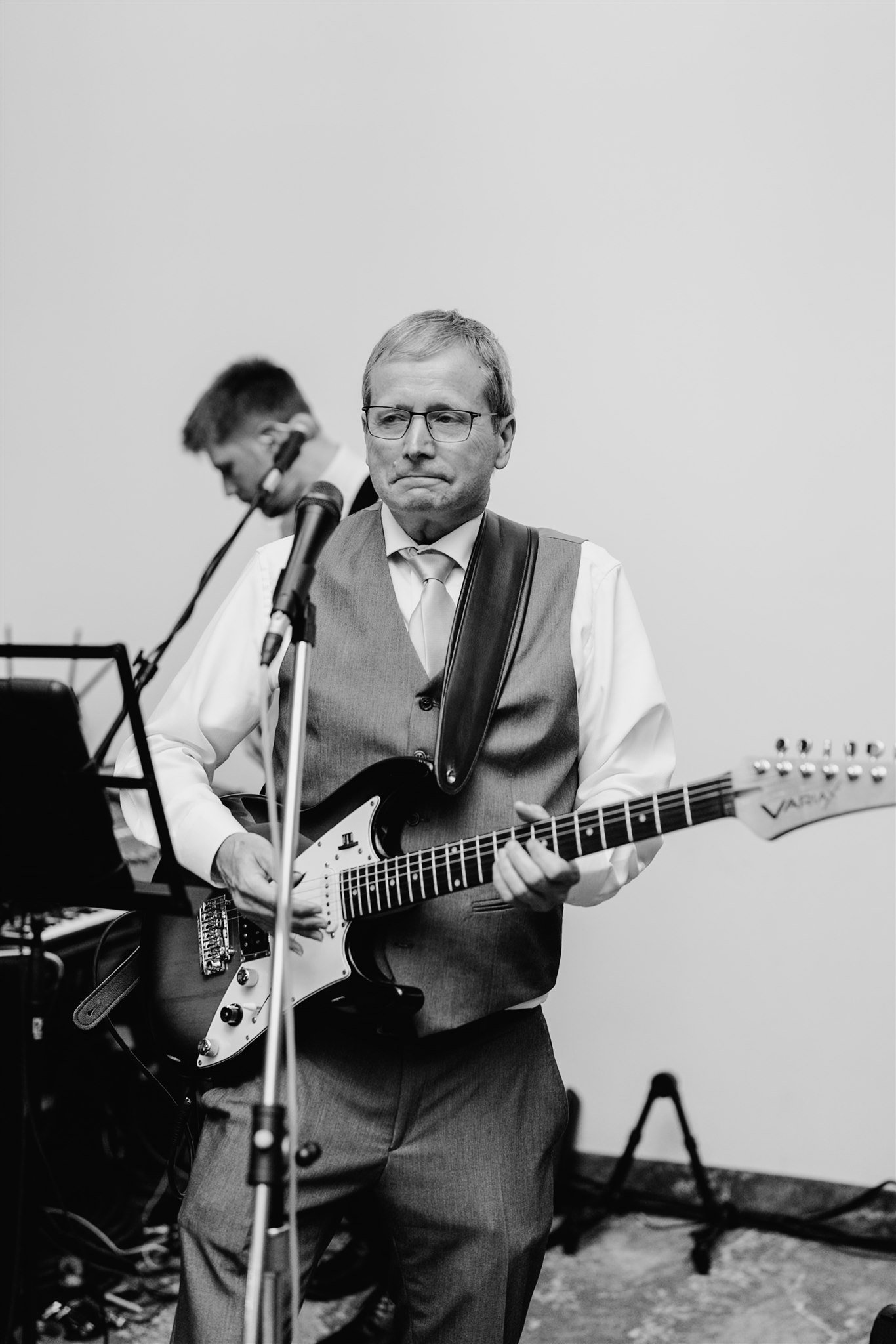 A man wearing glasses, a vest, and a tie playing an electric guitar with a serious expression, standing in front of a microphone, with another musician blurred in the background.
