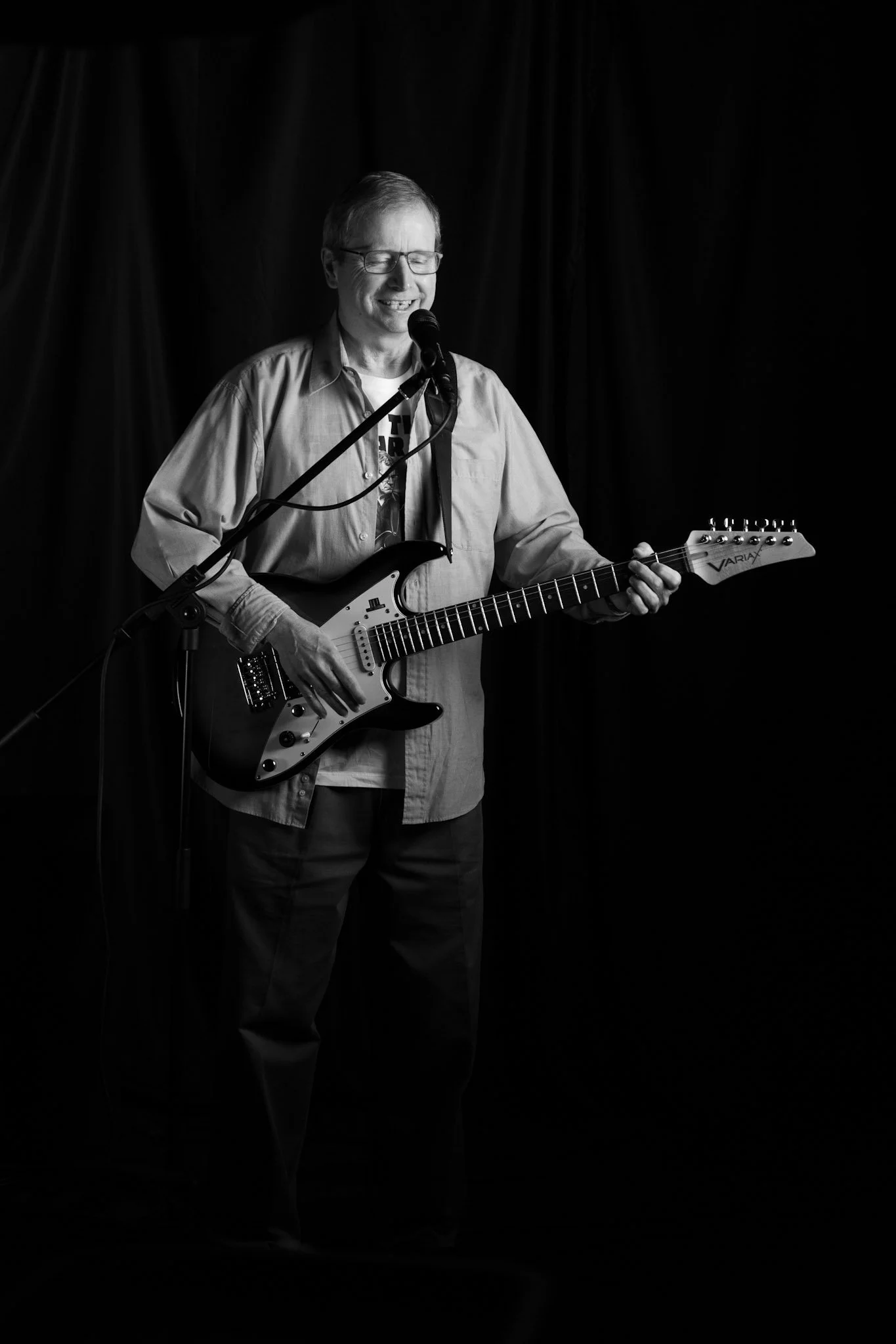 A man wearing glasses and casual clothing playing an electric guitar and singing into a microphone on stage with a black curtain background.