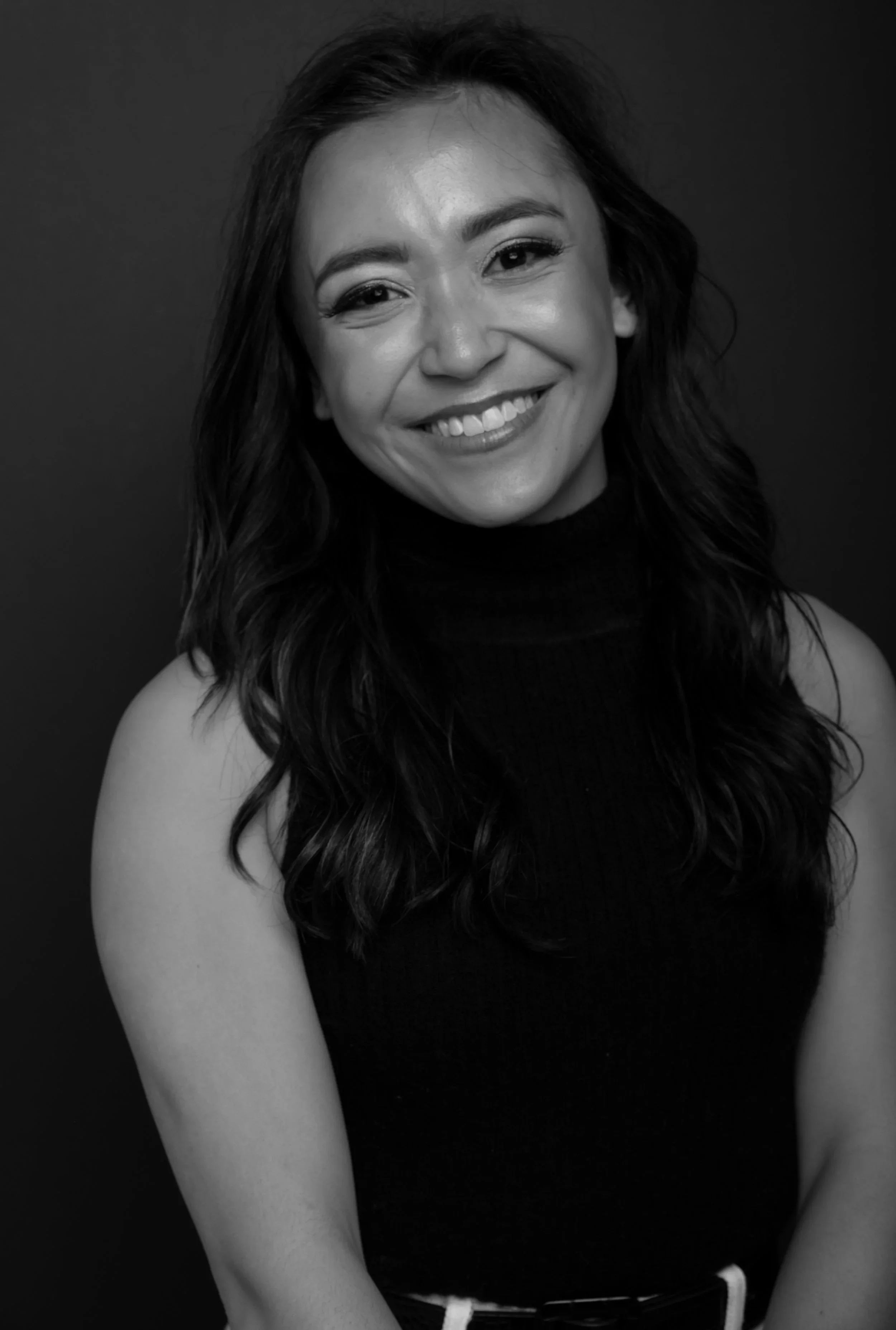 Black and white portrait of a woman with long wavy hair wearing a sleeveless turtleneck top, smiling against a dark background.