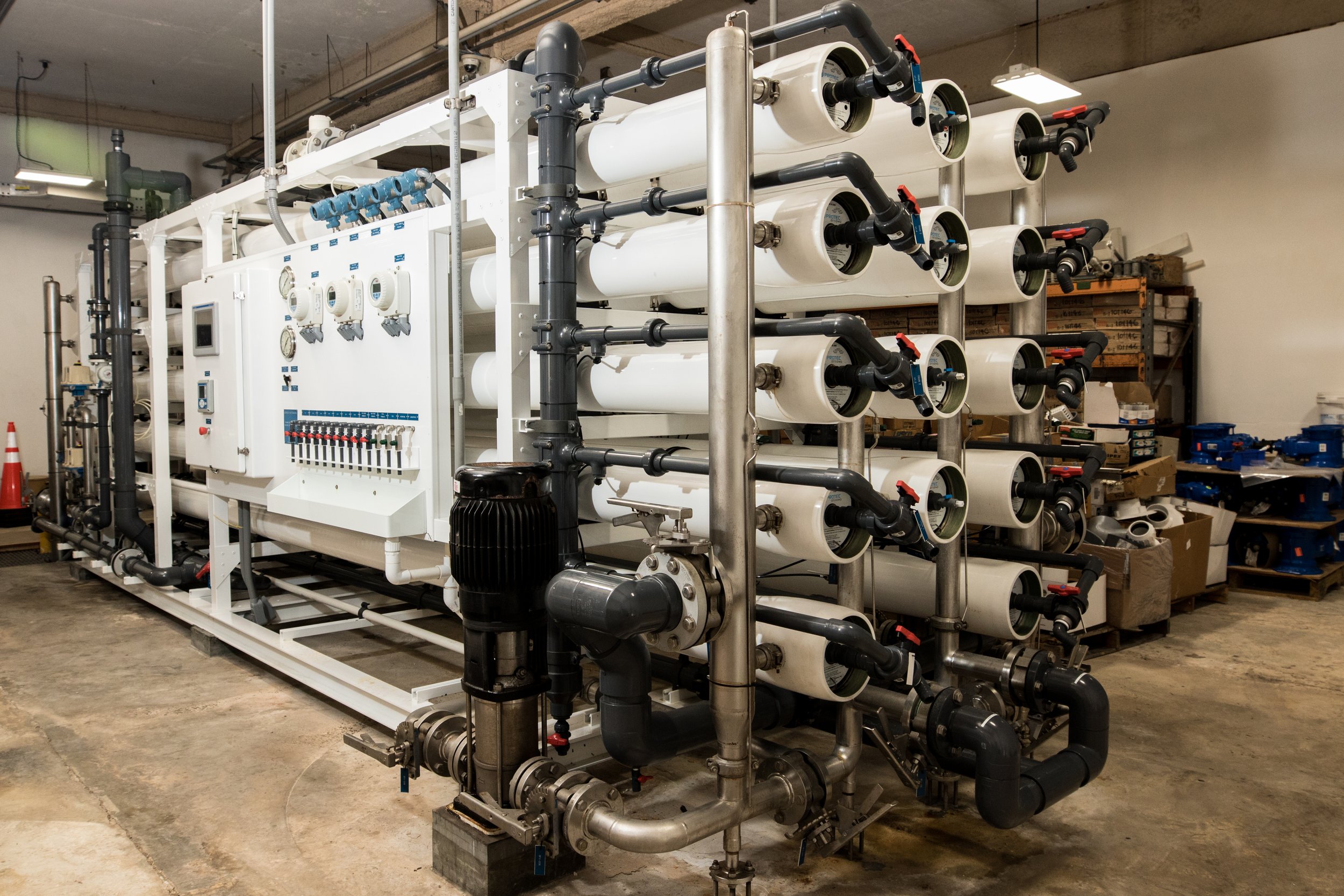 Industrial water filtration system with multiple white cylindrical filters, black pipes, and control panel, installed in a workshop or factory setting.