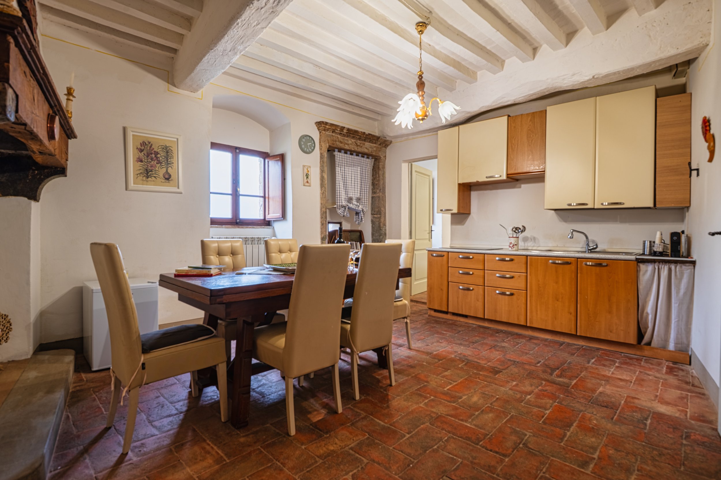 Interno di una cucina rustica con tavolo in legno e sedie beige, pareti bianche, pavimento in piastrelle rosse, finestre con persiane di legno, illuminazione con lampadario decorativo.