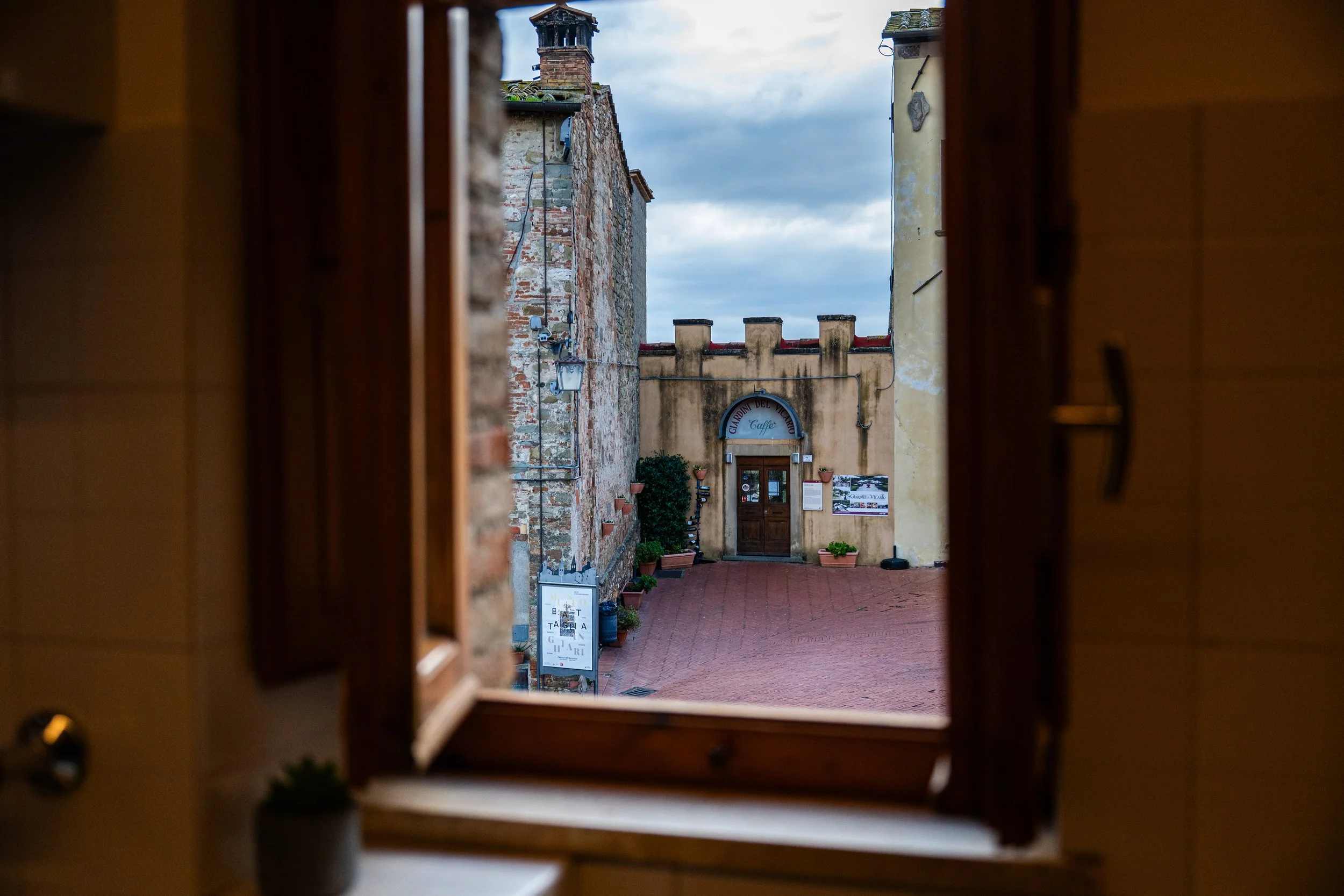 Vista di un cortile medievale attraverso una finestra aperta, con edifici in pietra e cartelli informativi.