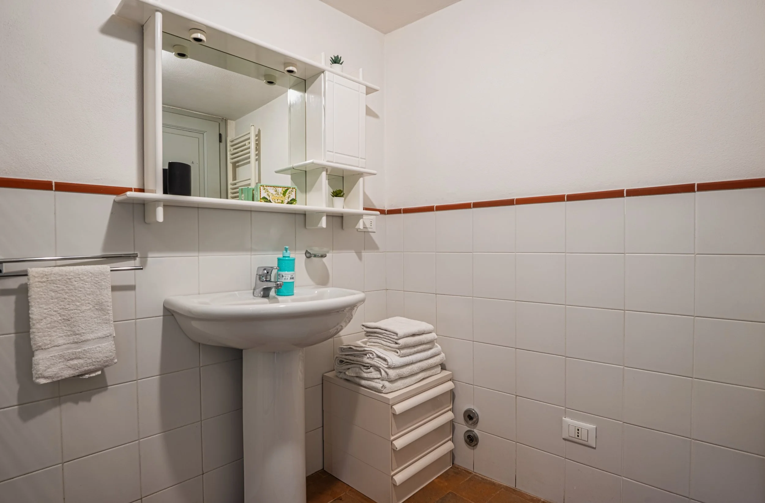 Bagno con lavabo, specchio, asciugamano e pile di asciugamani, mobile con cassetti, pareti bianche e piastrelle, presa elettrica e prese di ventilazione.