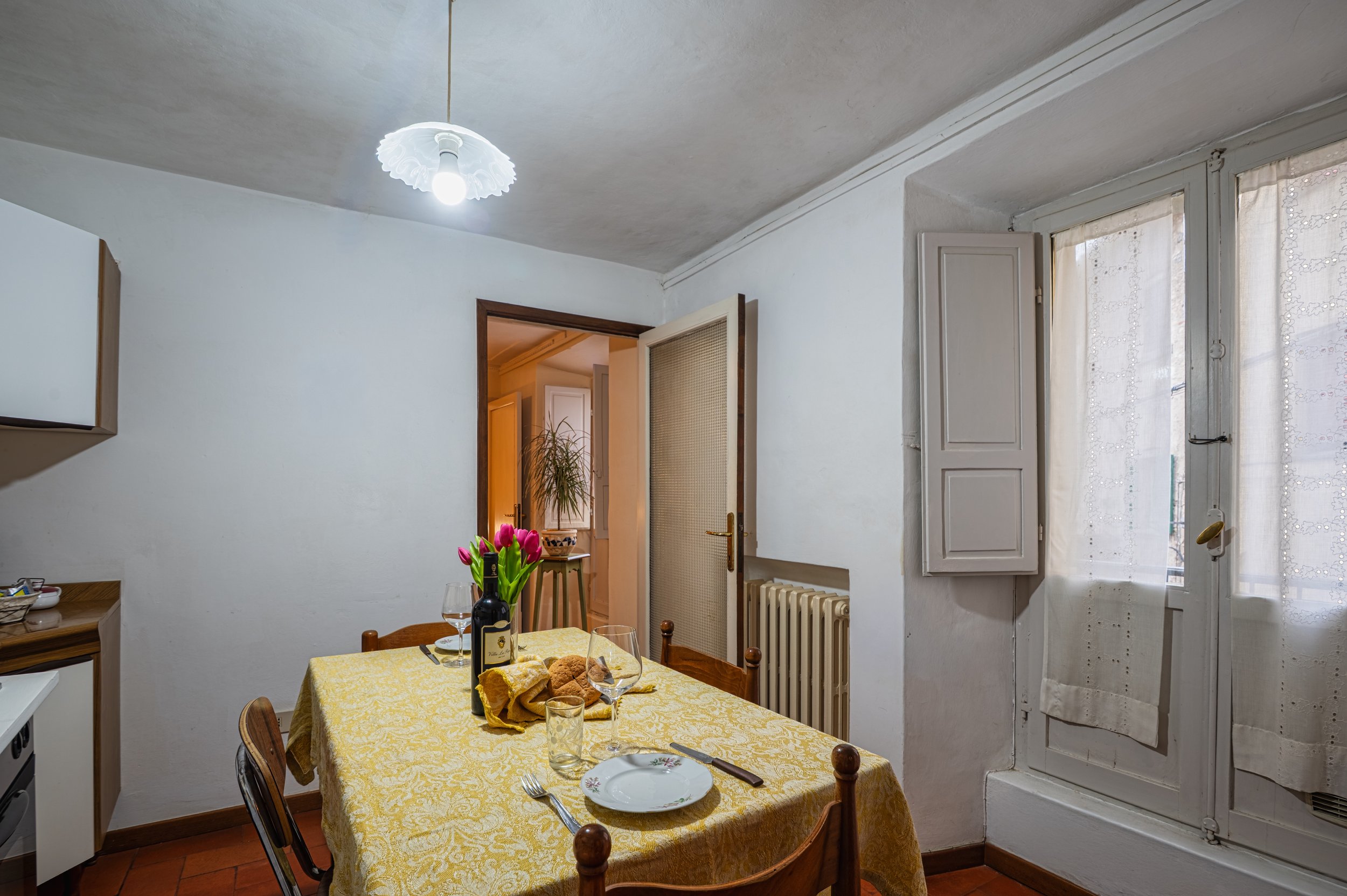 Sala da pranzo con tavolo rivestito di tovaglia gialla, piatti, bicchieri, coltelli, uno vaso con tulipani rosa, una bottiglia di vino, e pane. Finestra con tende chiare. Porta aperta che porta a un altro ambiente con piante. Illuminazione artificial