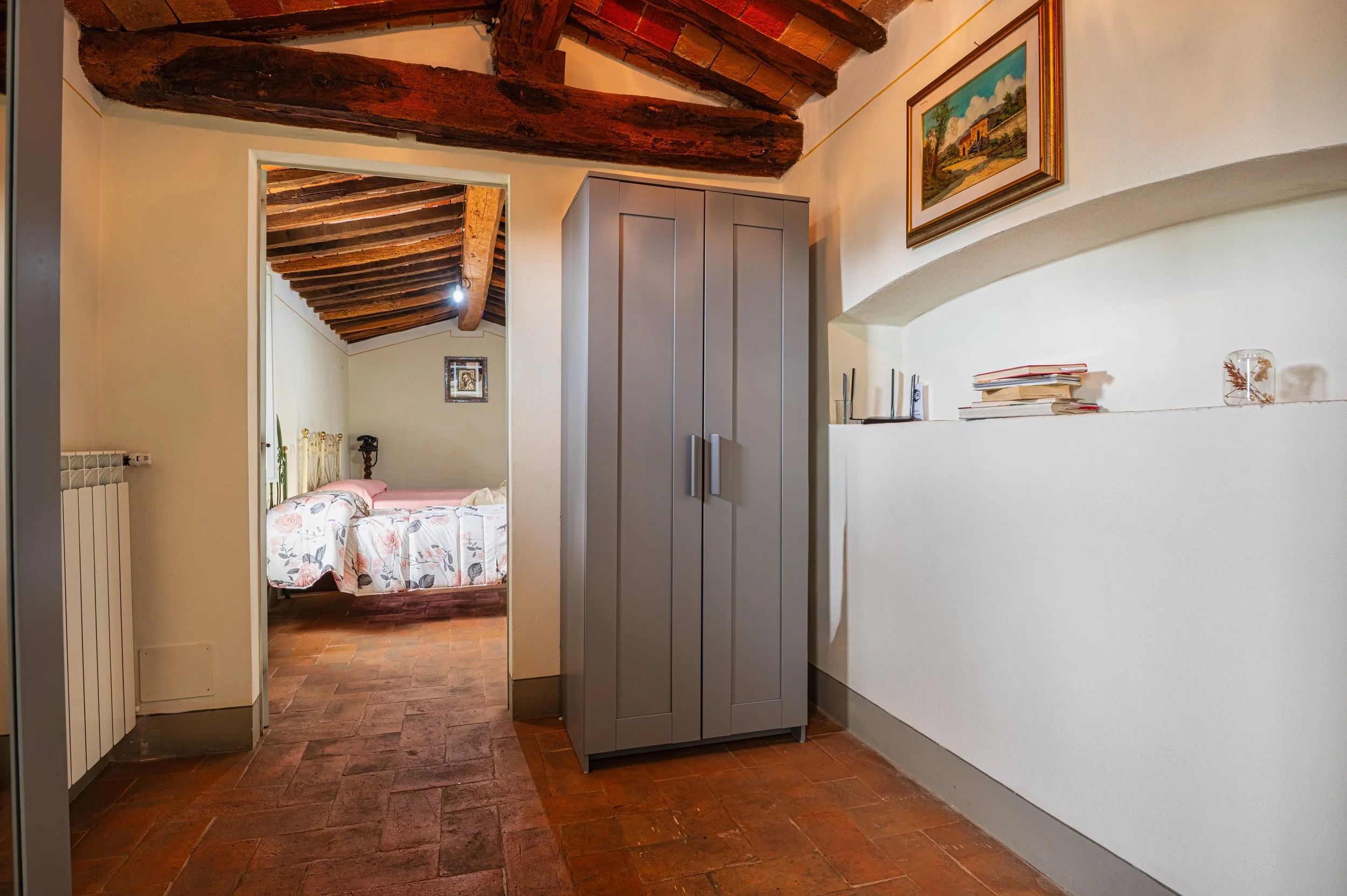 Interno di una casa con soffitto in legno a vista, pareti bianche, un armadio grigio, e una nicchia con libri e decorazioni. Si intravede una camera da letto con un letto in ferro battuto e una parete decorata con un quadro.