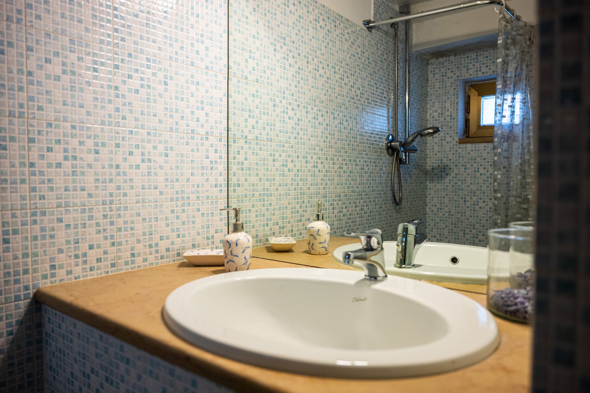 Vasche da bagno con rubinetto su un piano di legno, specchio grande, saponi e bicchieri con decorazioni, pareti rivestite di piastrelle celesti con piccoli mosaici, doccia con soffione a soffitto e piccolo finestrino