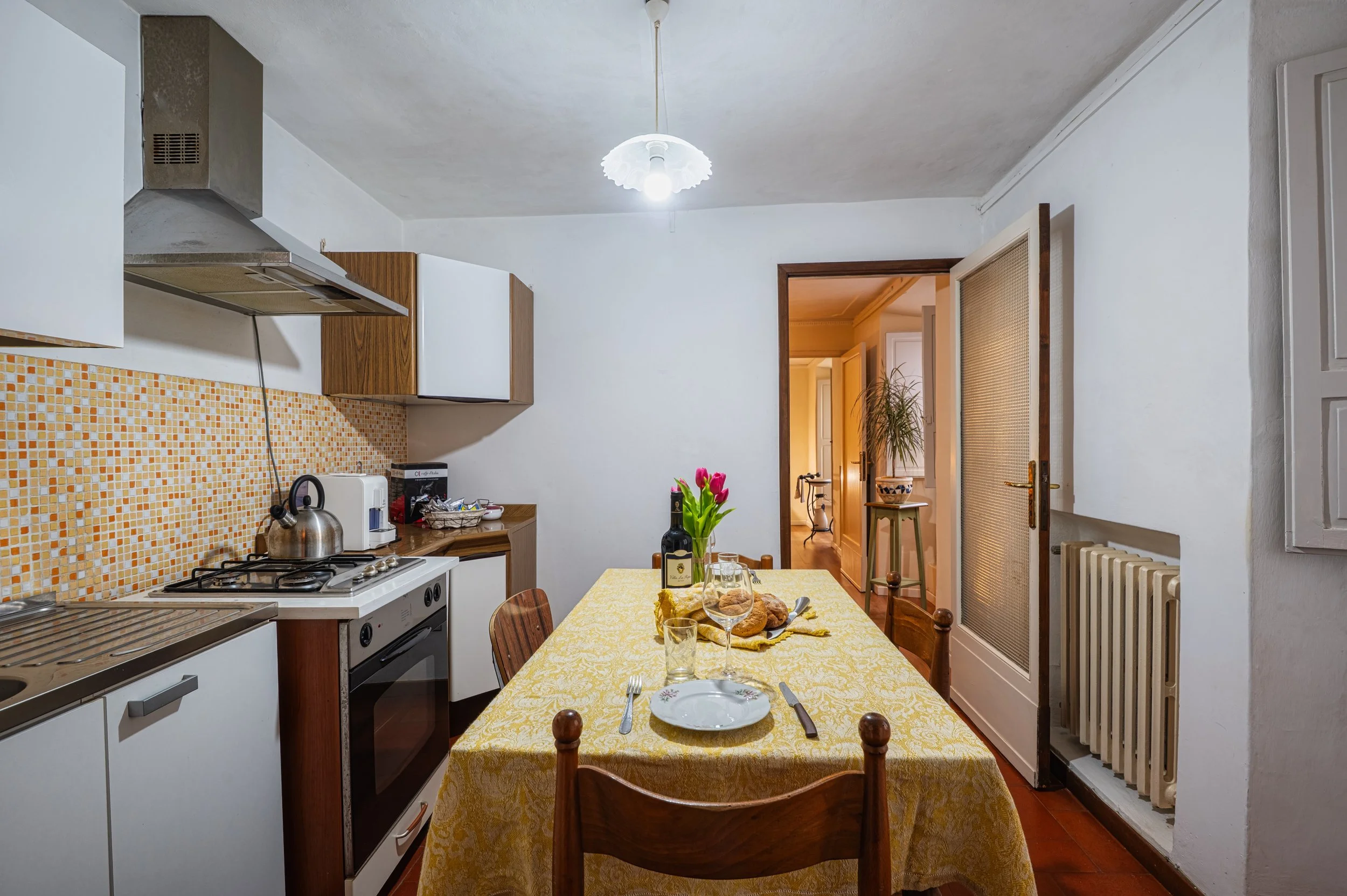 Sala da pranzo con tavolo coperto da tovaglia gialla, sedie di legno, piatto, bicchieri, posate, bottiglia di vino, vase con fiori, e pane.