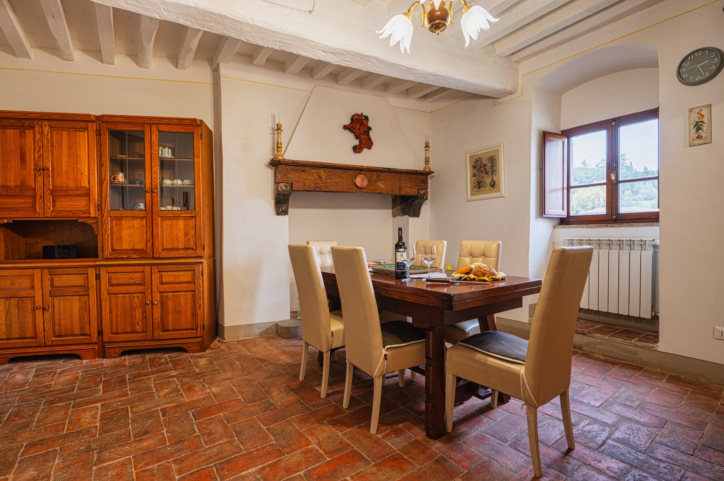 Room with wooden cabinet, fireplace mantel with decorative items, dining table set for meal with wine, plates, glasses, and bread; tiled floor, white walls, window with open shutters, and ceiling with wooden beams.