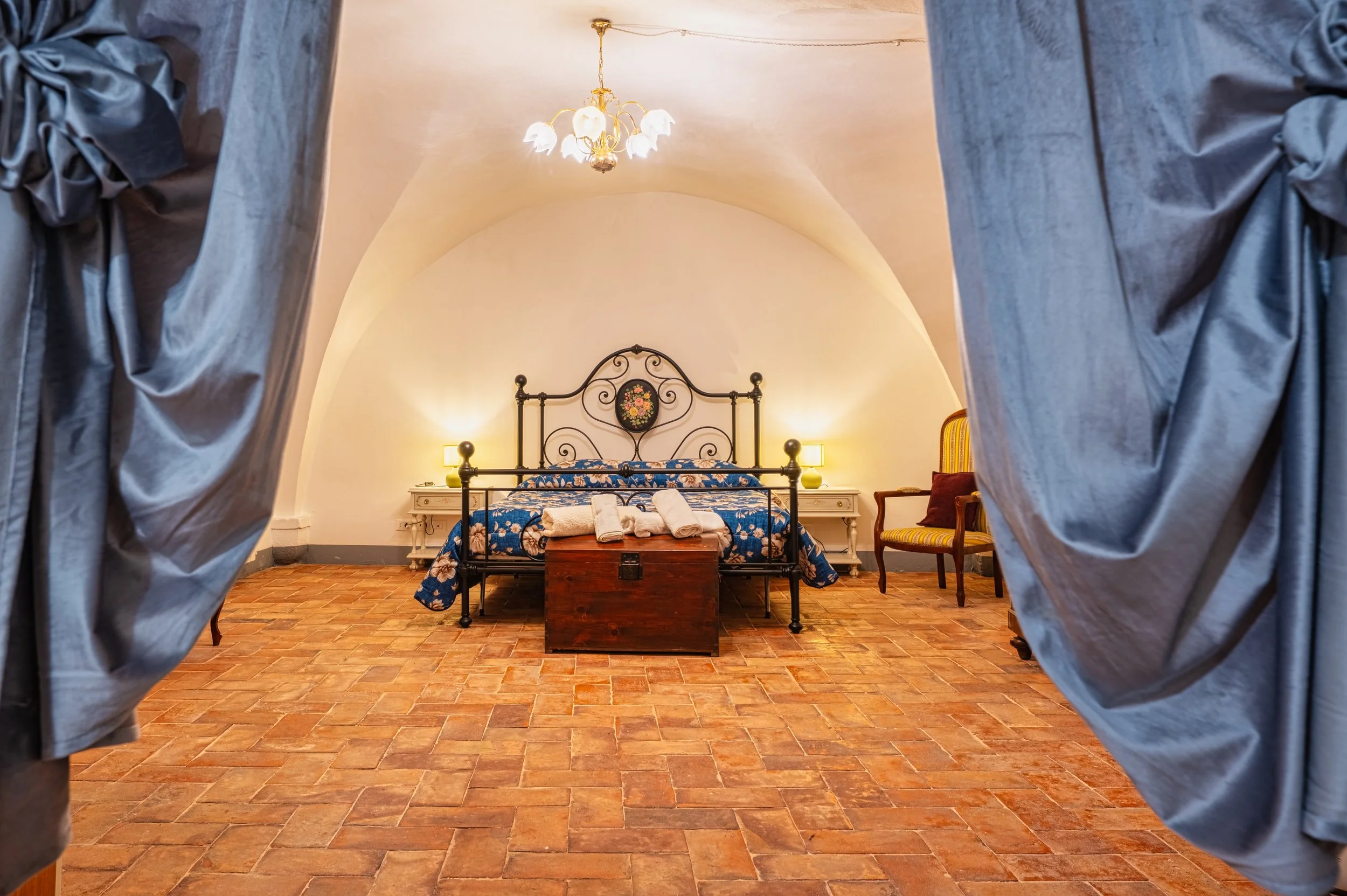 Vista di una camera da letto con pareti bianche, un letto con testiera in ferro battuto e copriletto blu con motivi floreali, un tappeto di pietra e una sedia con schienale in legno e tessuto giallo. I tendaggi di seta blu sono aperti, rivelando l'interno della stanza.