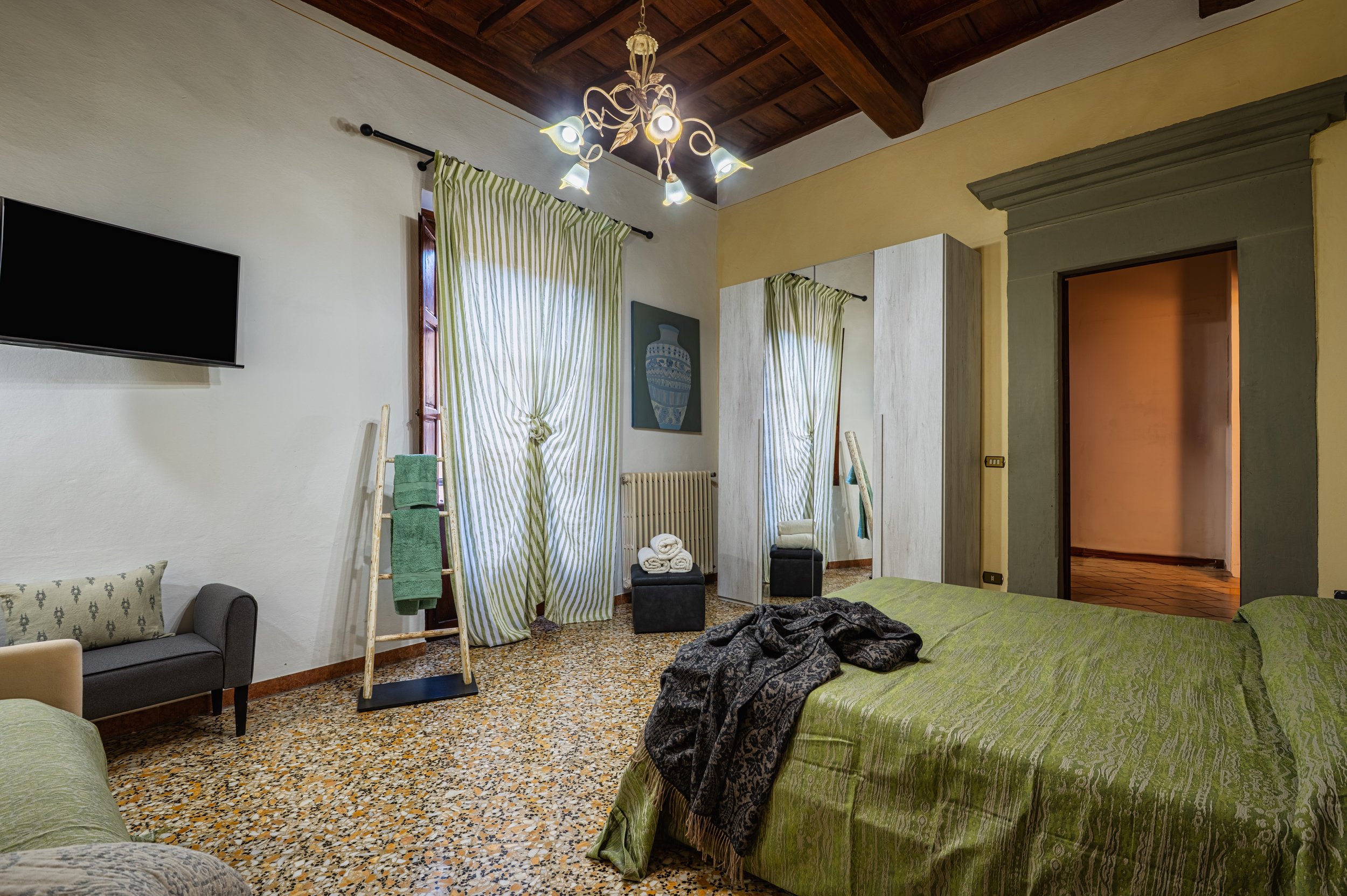 Camera da letto con pareti di colore chiaro, letto con coperta verde, armadio con specchio, finestra con tende a righe verdi e bianche, lampadario a soffitto, televisore a parete, porta aperta che mostra un'altra stanza, e vari dettagli di arredament