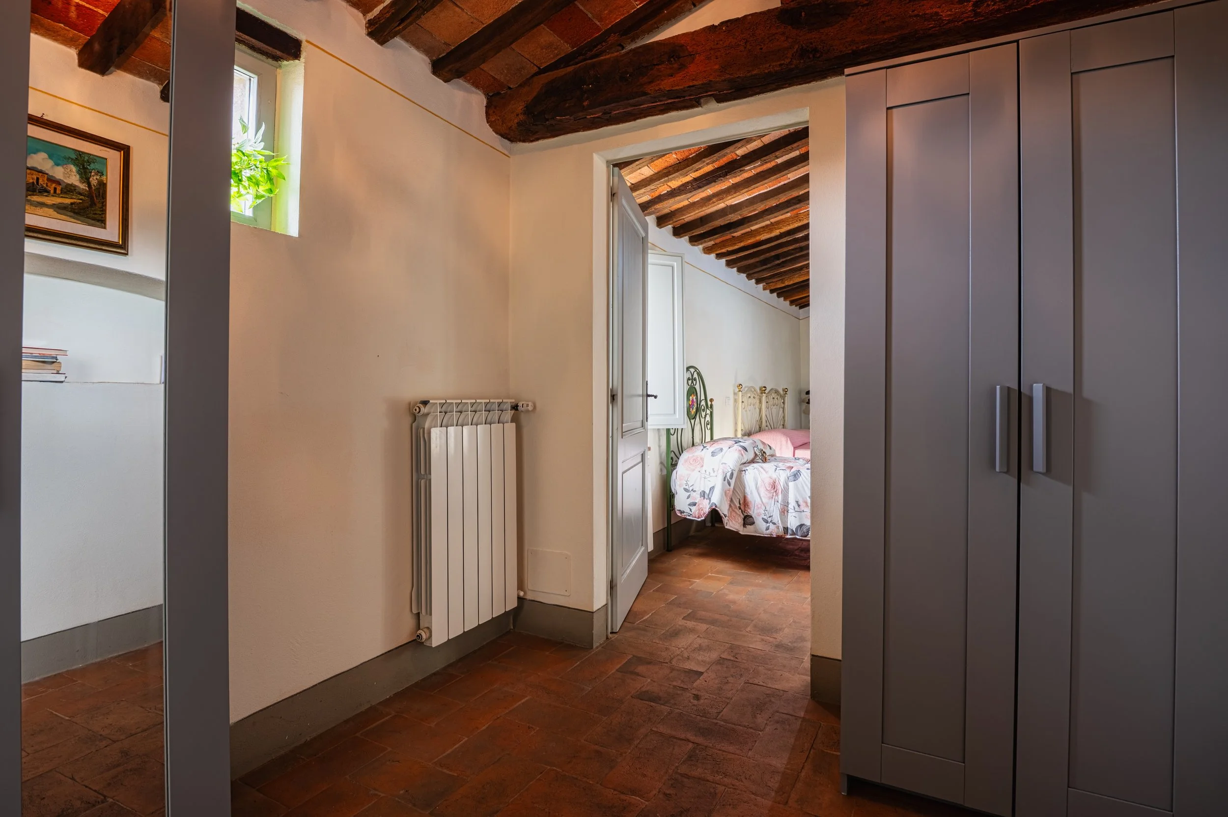 Vista di una camera da letto attraverso un ingresso, con soffitto in legno e pareti bianche, arredi moderni, letto con copriletto floreale e una finestra con pianta.