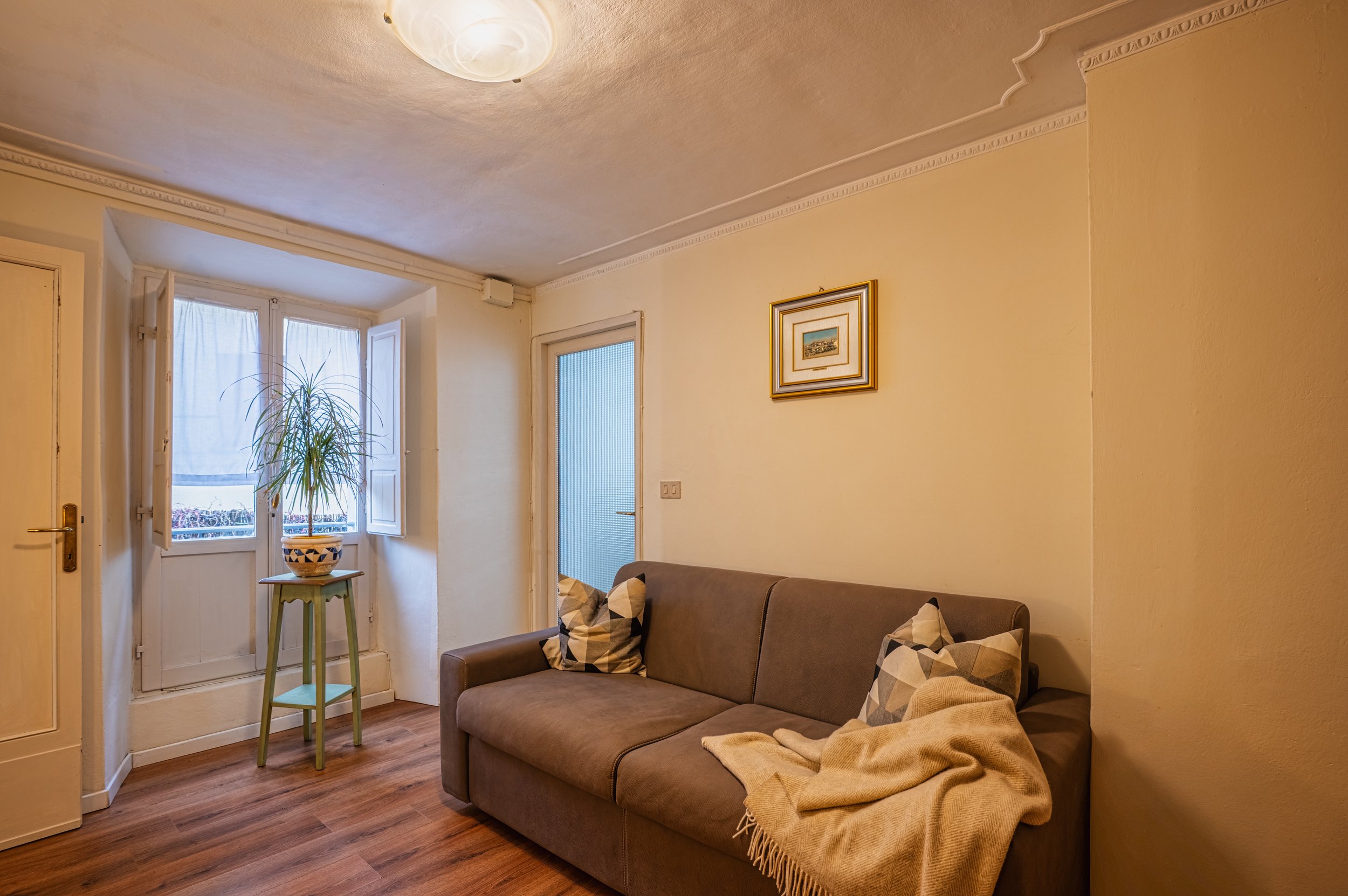 Soggiorno con divano marrone, cuscini geometrici, coperta beige, pareti gialle, quadro sopra il divano, finestra con piante in vaso, pavimento in legno.