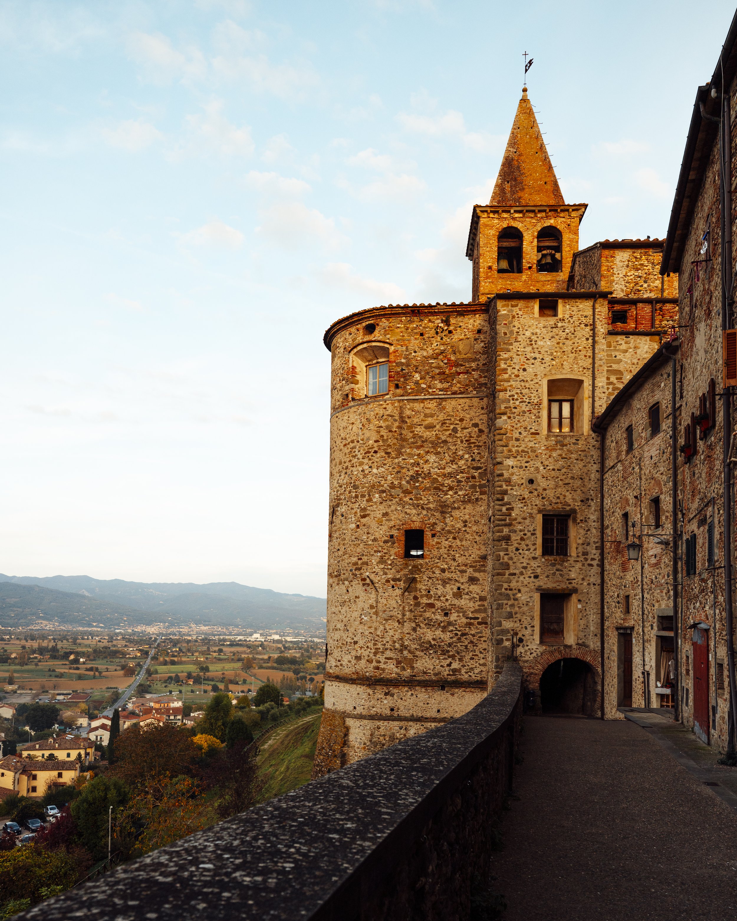 Torri medievali di pietra in un villaggio, con una strada che le attraversa, e un paesaggio rurale con campi e montagne sullo sfondo