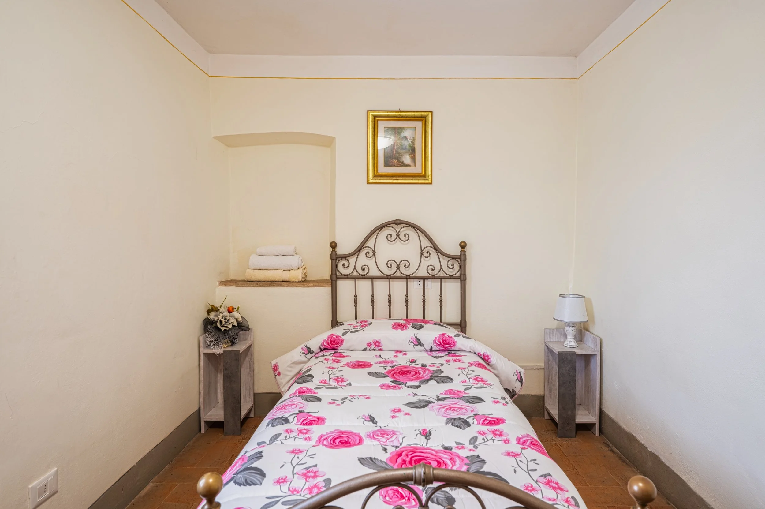 Camera da letto con letto singolo, copriletto con motivo di rose rosa e foglie grigie, due comodini con lampada e fiori, quadri sopra la testata del letto, coperte piegate su una nicchia nel muro.