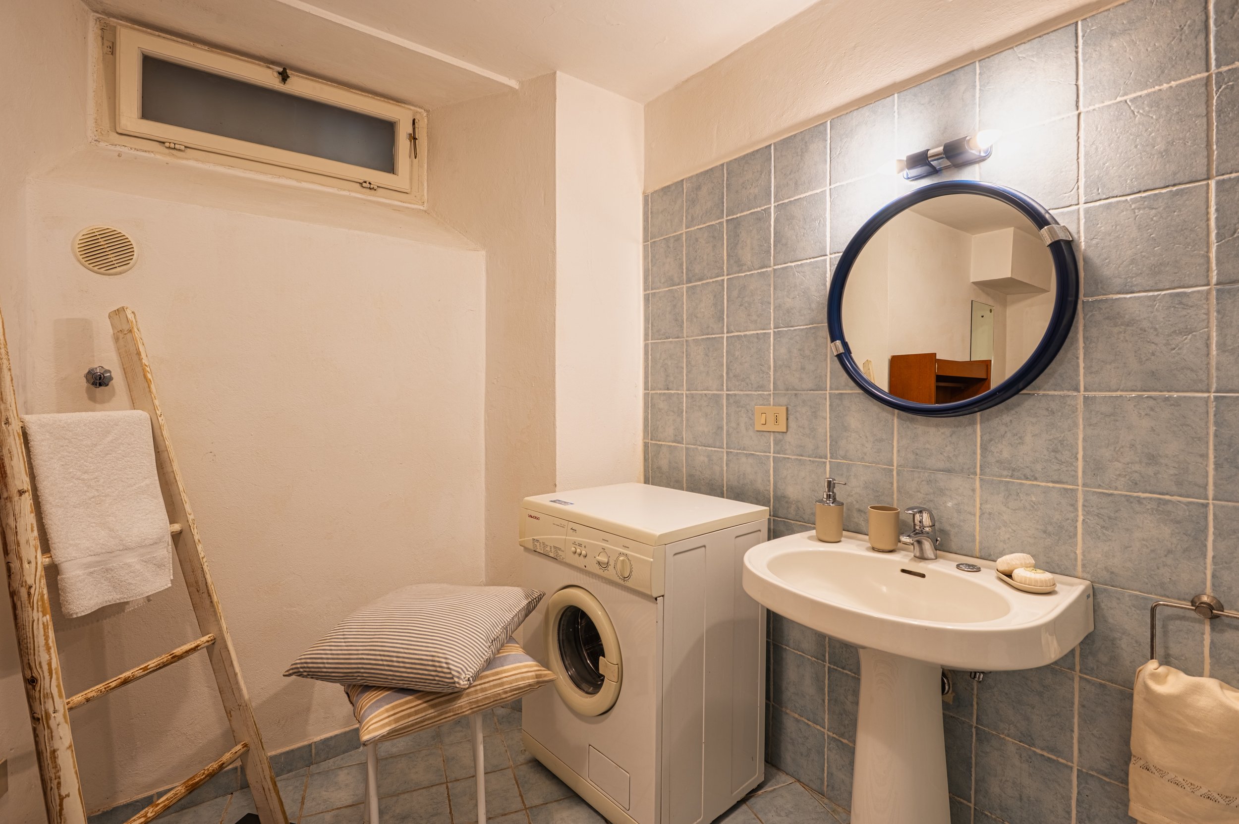 Bagno con lavatrice, lavabo e specchio rotondo, pareti rivestite di piastrelle azzurre, finestra piccola, scala di legno con asciugamano, sedile con cuscini