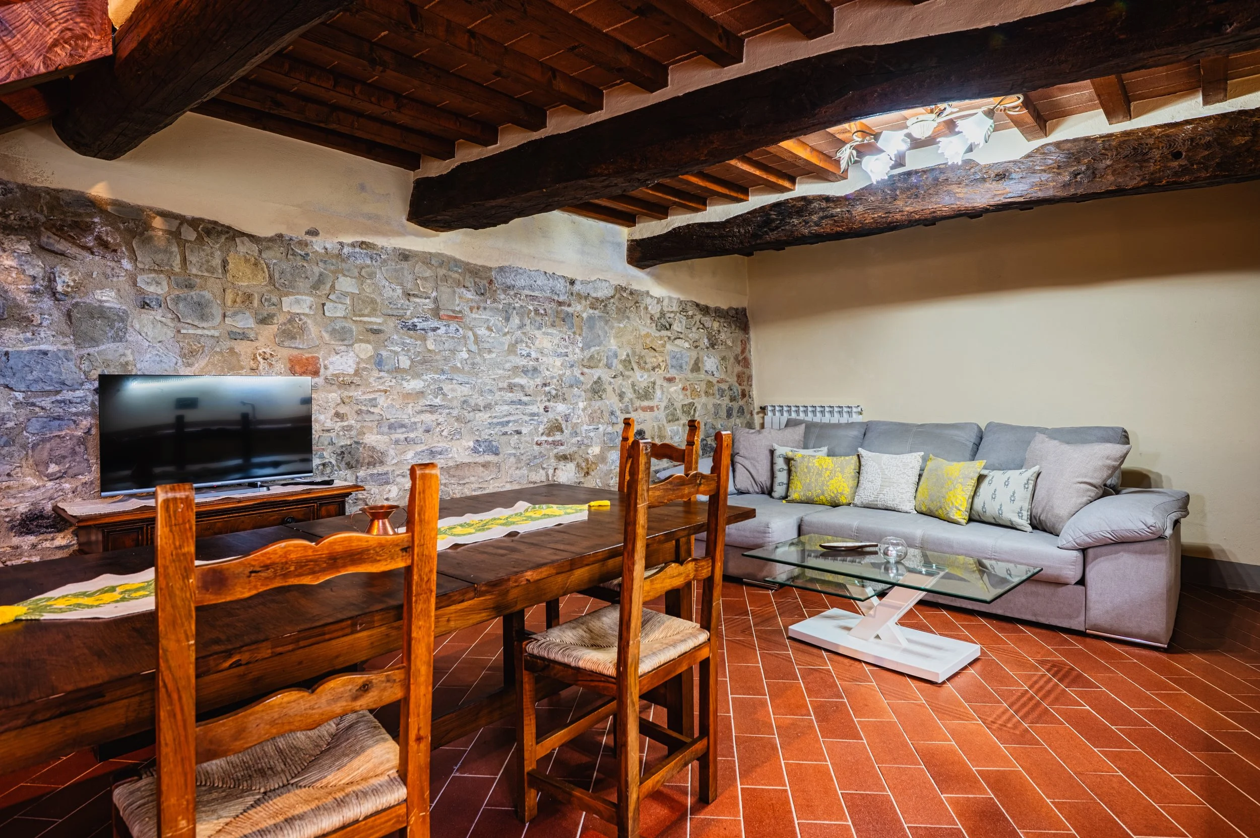 Soggiorno con parete in pietra, divano grigio, tavolino in vetro, sedie in legno, soffitto con travi di legno e lampadario