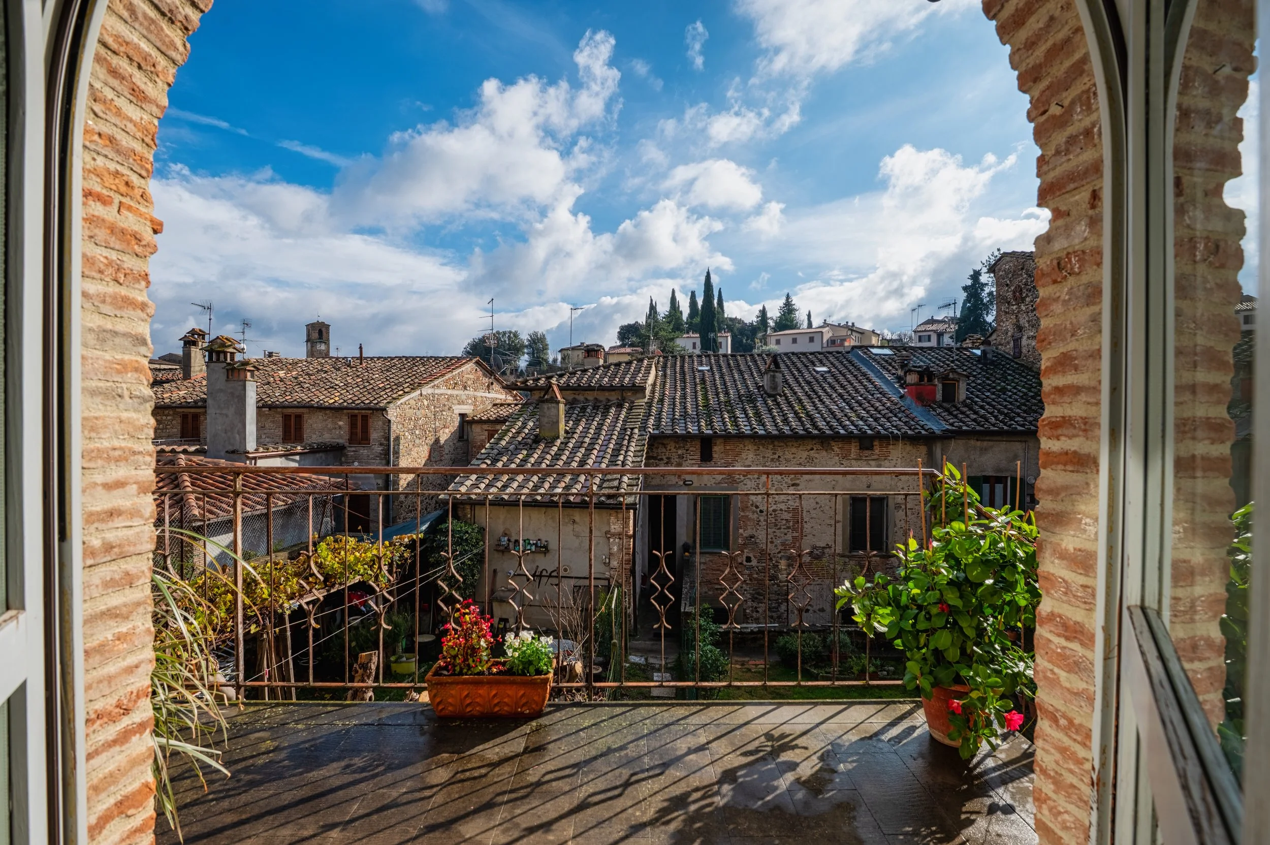 Vista da una finestra di un terrazzo con fiori in vasi, guarda su un tetto di ceramica e un cielo con nuvole