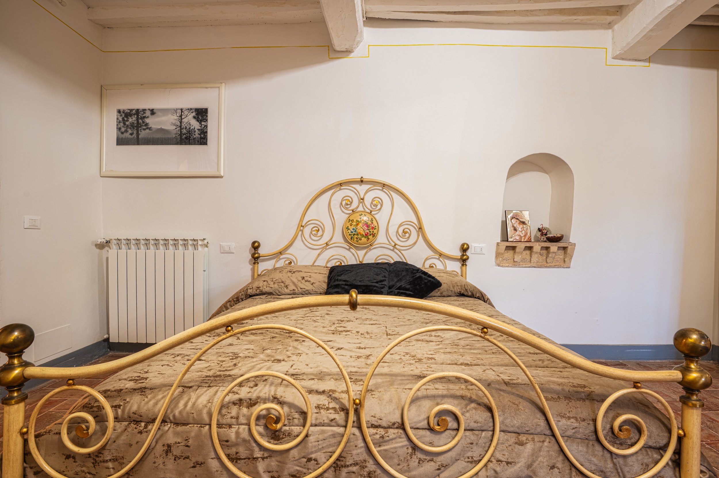 Letto matrimoniale con testiera di metallo dorato, copriletto beige con motivo floreale, cuscini neri e marroni, pareti bianche, quadro con paesaggio, nicchia con oggetti e cornice nera, termosifone bianco, pavimento in terracotta.