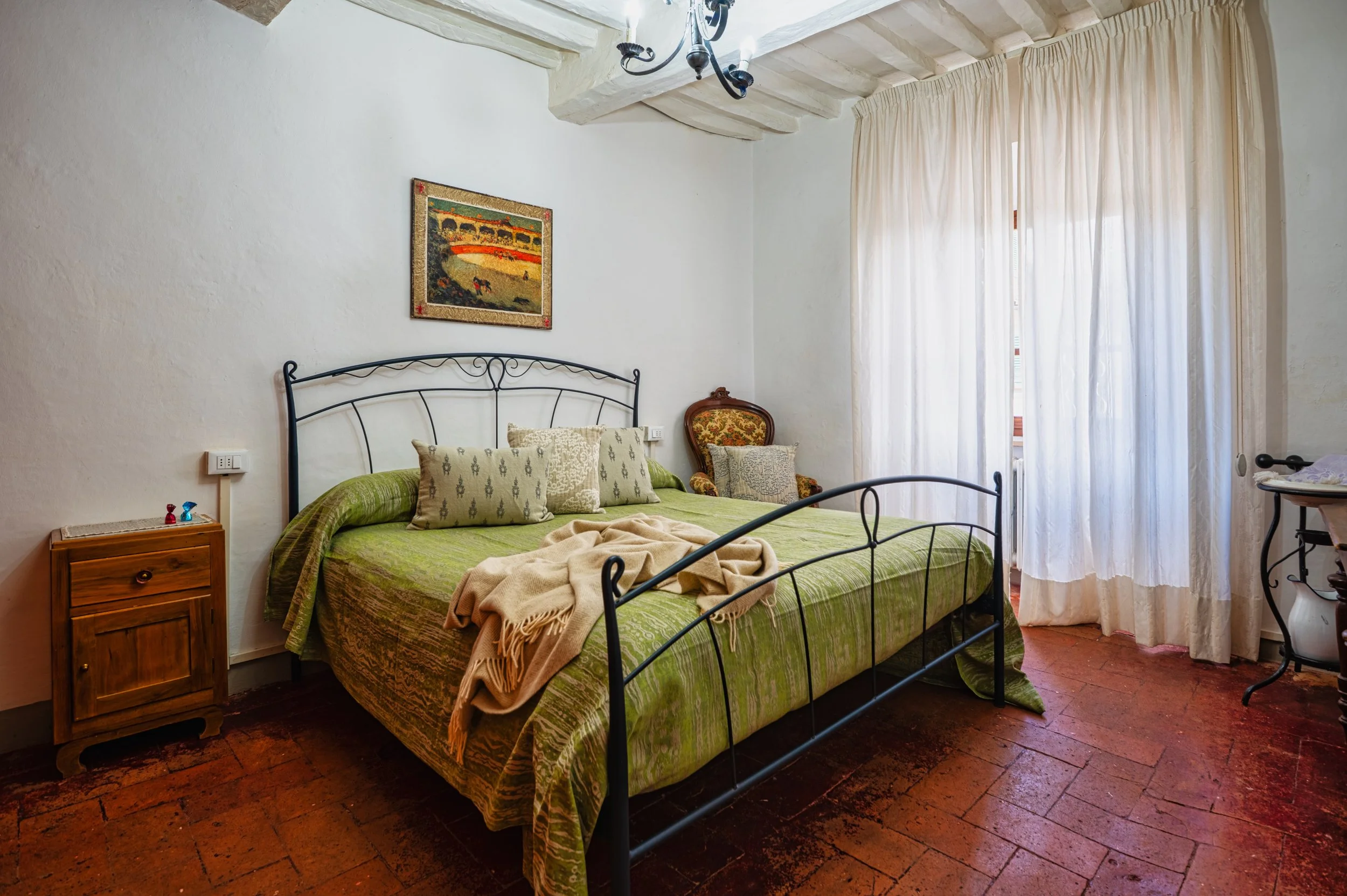 Stanza da letto con letto matrimoniale con copriletto verde, fra alcuni cuscini dai motivi vari, una macchia di coperta beige, una sedia di stoffa con motivo floreale e una finestra con tende bianche.