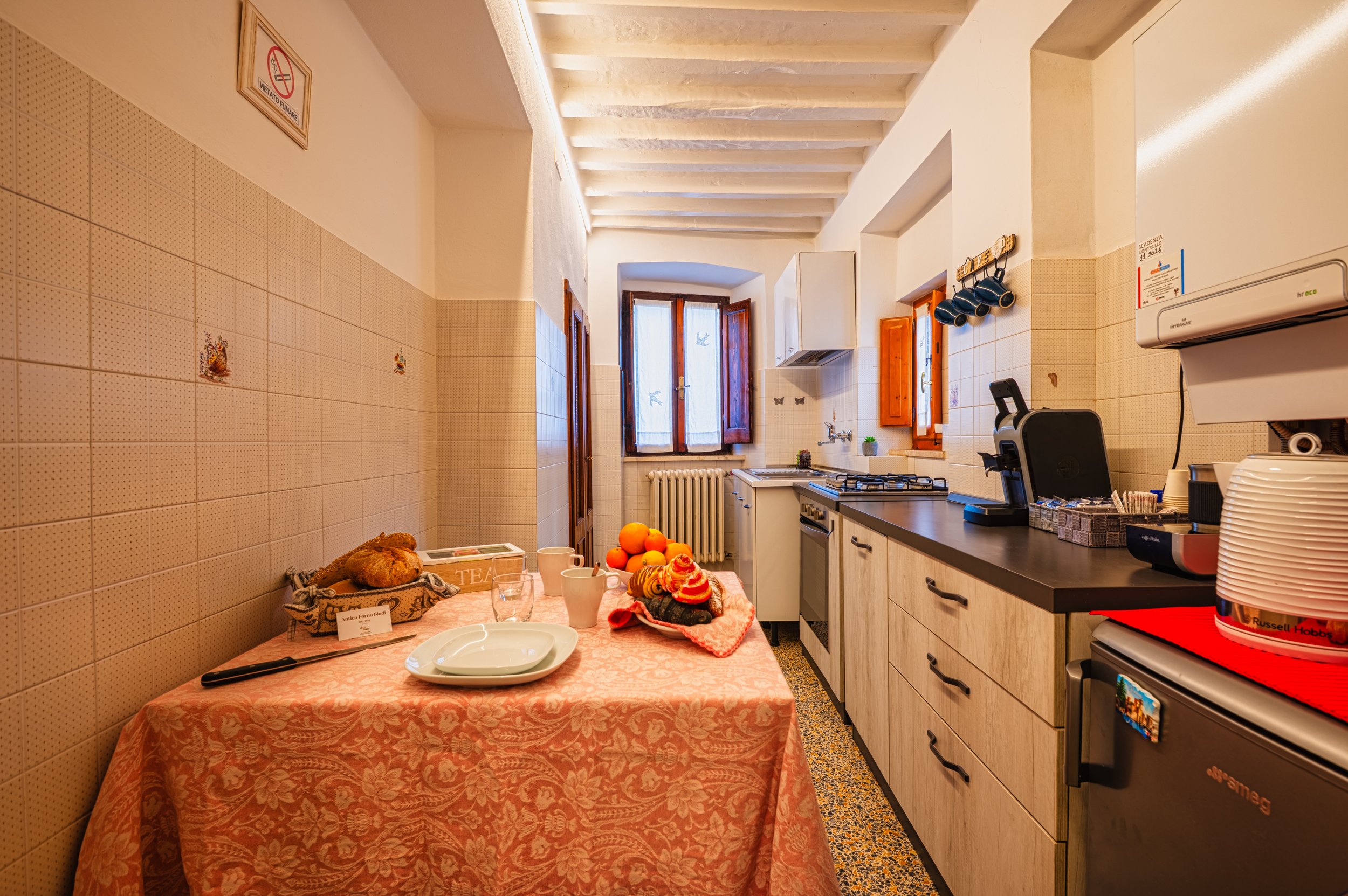 Cucina rustica con tavolo apparecchiato, cucina a gas, frigorifero, finestre e pareti rivestite di piastrelle beige con decorazioni.