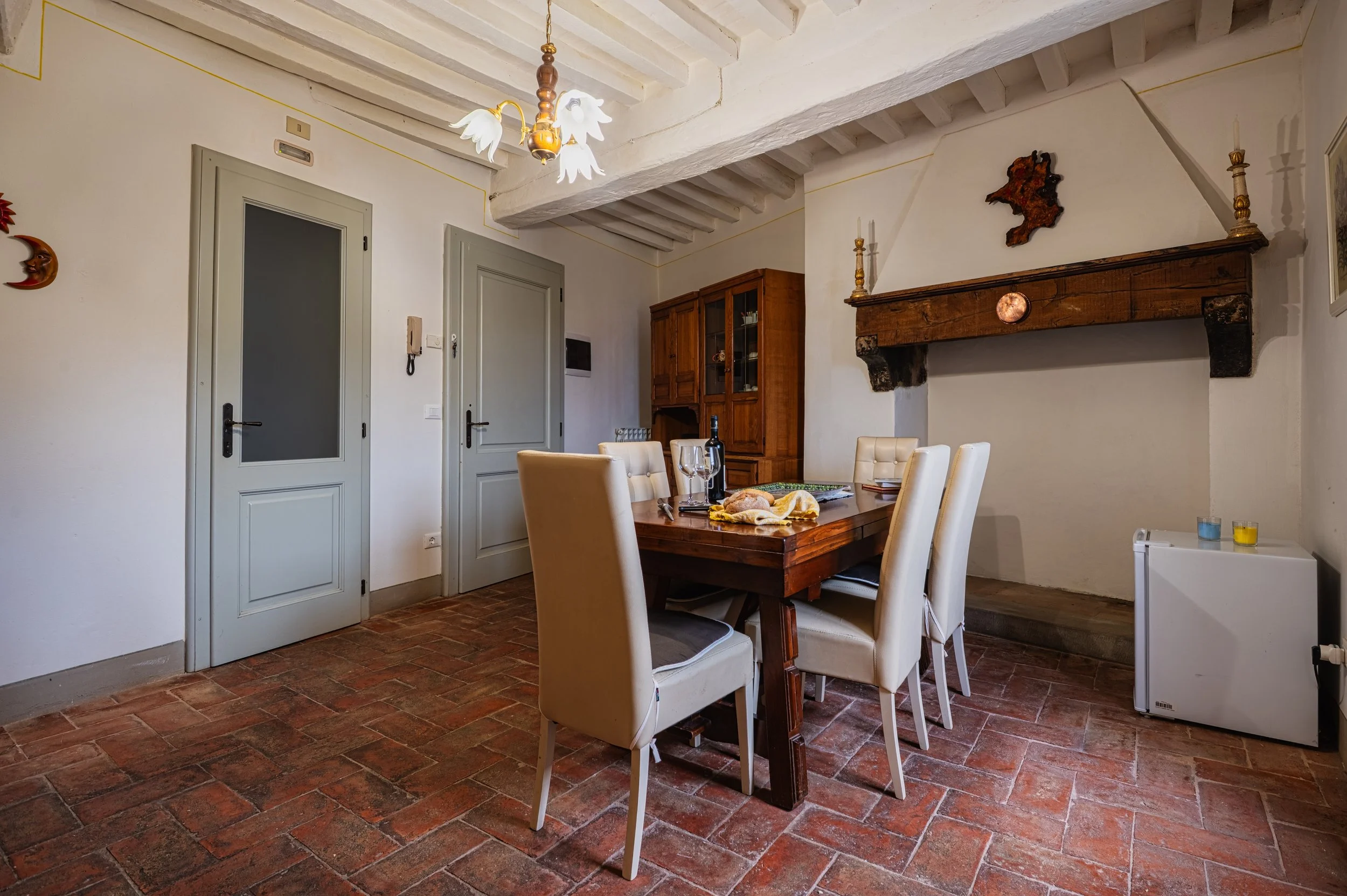 Una sala da pranzo con tavolo in legno e sedie imbottite color crema. Sul tavolo ci sono un'asse di pane e una bottiglia di vino, con alcuni bicchieri. Sul muro, un camino in mattoni e decorazioni, tra cui una scultura astratta e candelabri. Le paret