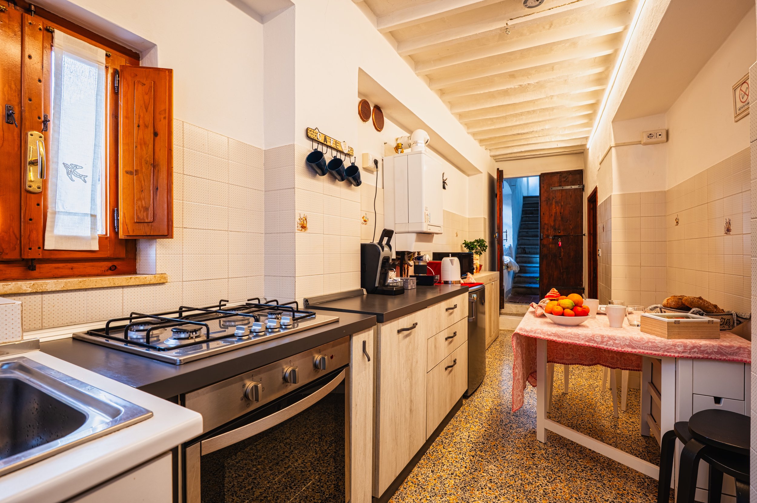 Cucina rustica con pareti bianche e soffitto di legno, piana di lavoro con elettrodomestici e stoviglie, tavolo con frutta e pane.