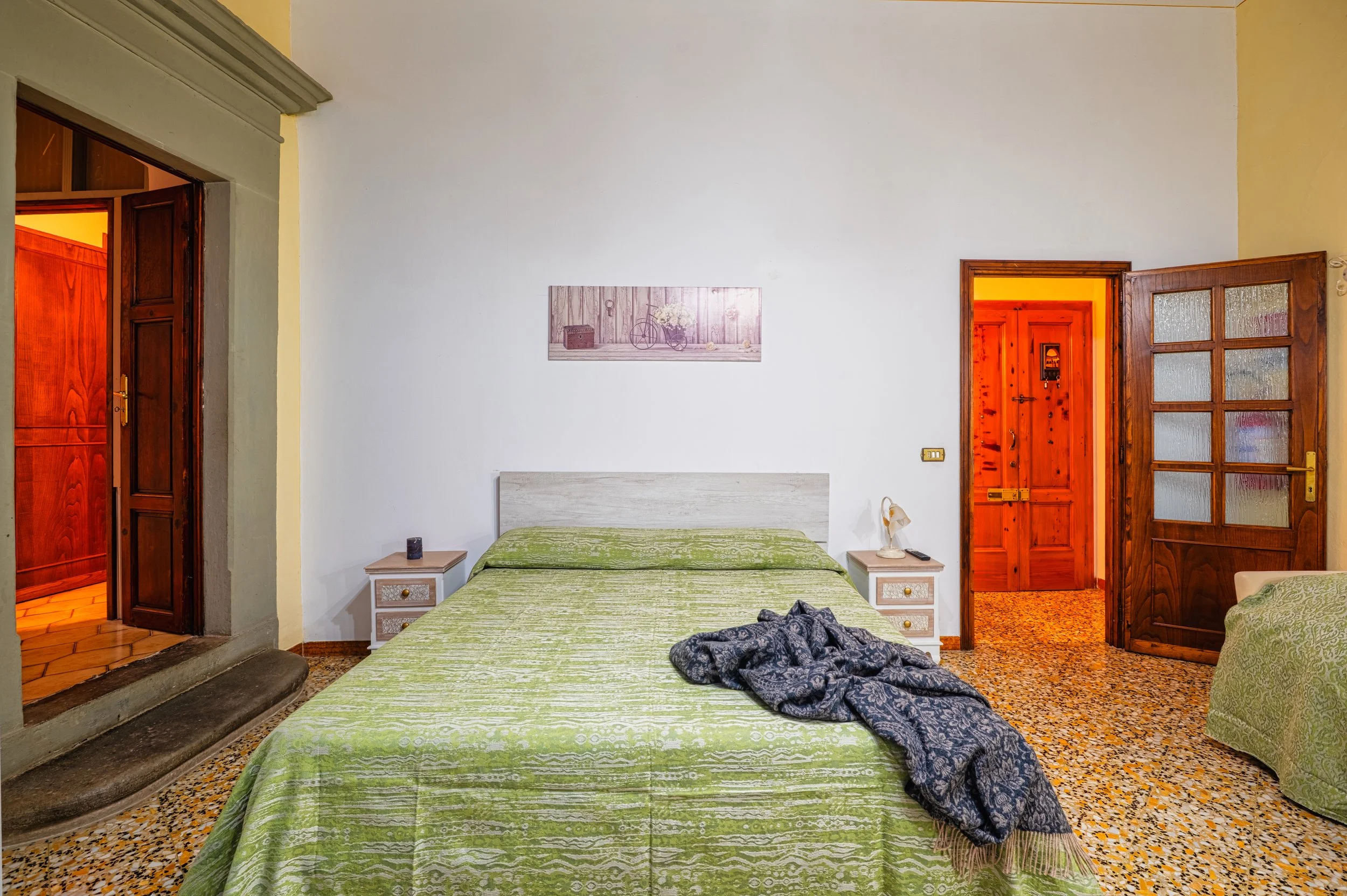 Camera da letto con letto matrimoniale, due comodini con lampada e telefono, parete bianca con quadro, porta di ingresso in legno, pavimento in marmo colorato.