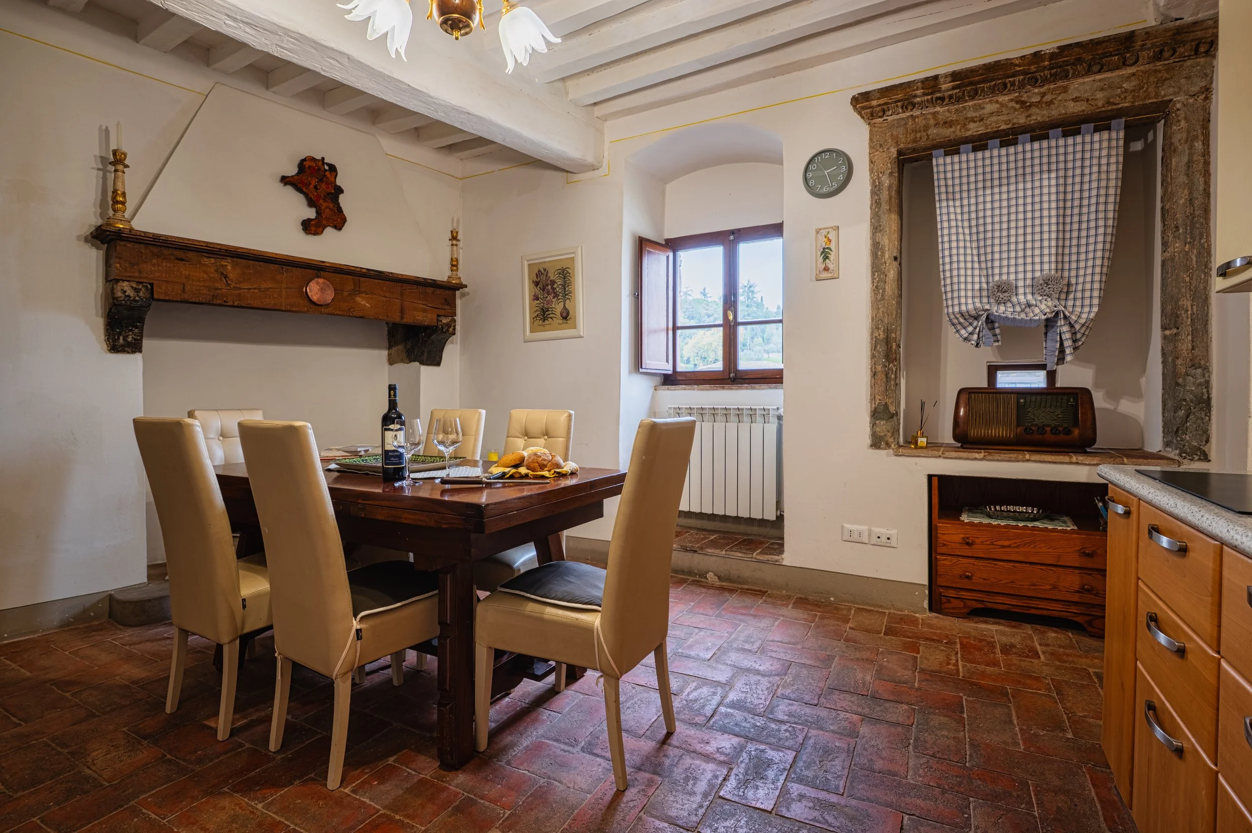 Sala da pranzo con tavolo in legno, sei sedie imbottite color crema, alimenti e bicchieri, camino antico in pietra, finestra con vista alla natura, pareti in pietra e legno, tappeti in mattonelle di terracotta.