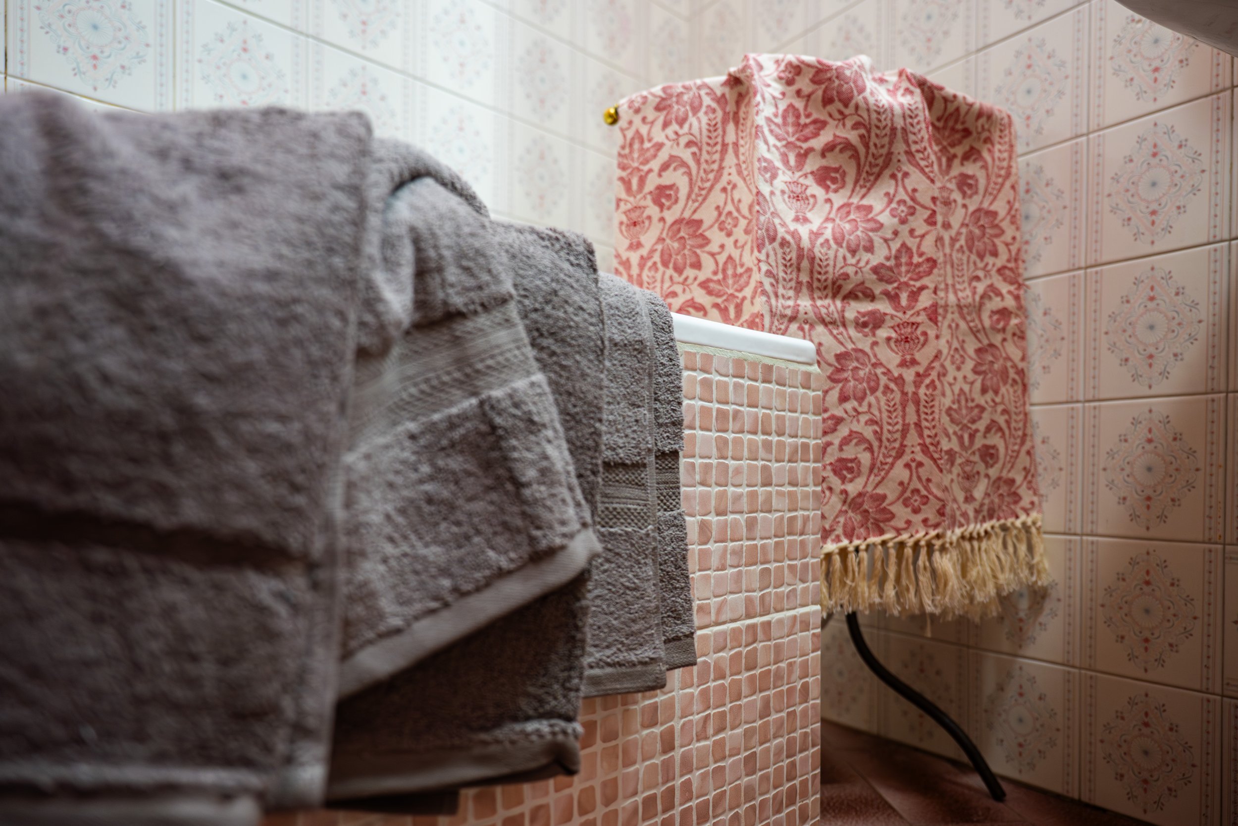 Asciuga asciugamani grigi appoggiati in bagno su un mobile rivestito di piastrelle rosa, con una lavatrice coperta da un panno decorato di colore rosa con motivi floreali e frange.