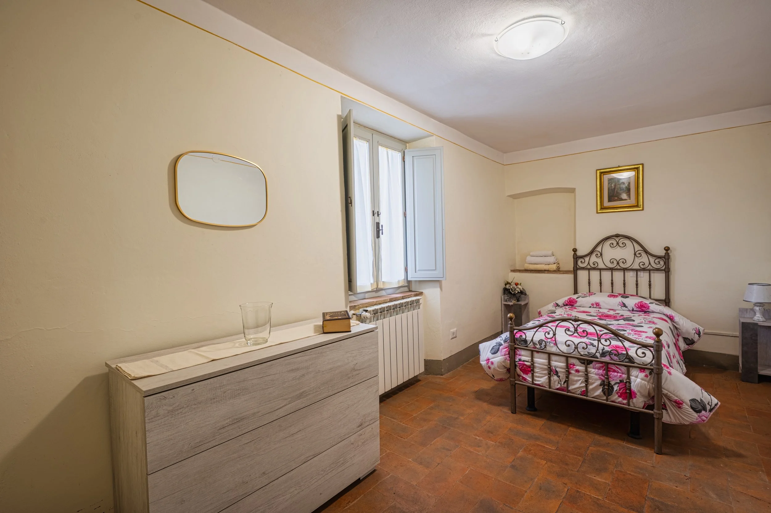 Camera da letto con letto a ferro battuto, coperta floreale, finestra con persiane bianche, piccolo quadro appeso alla parete, comodino con lampada, e finestra aperta con tende bianche.