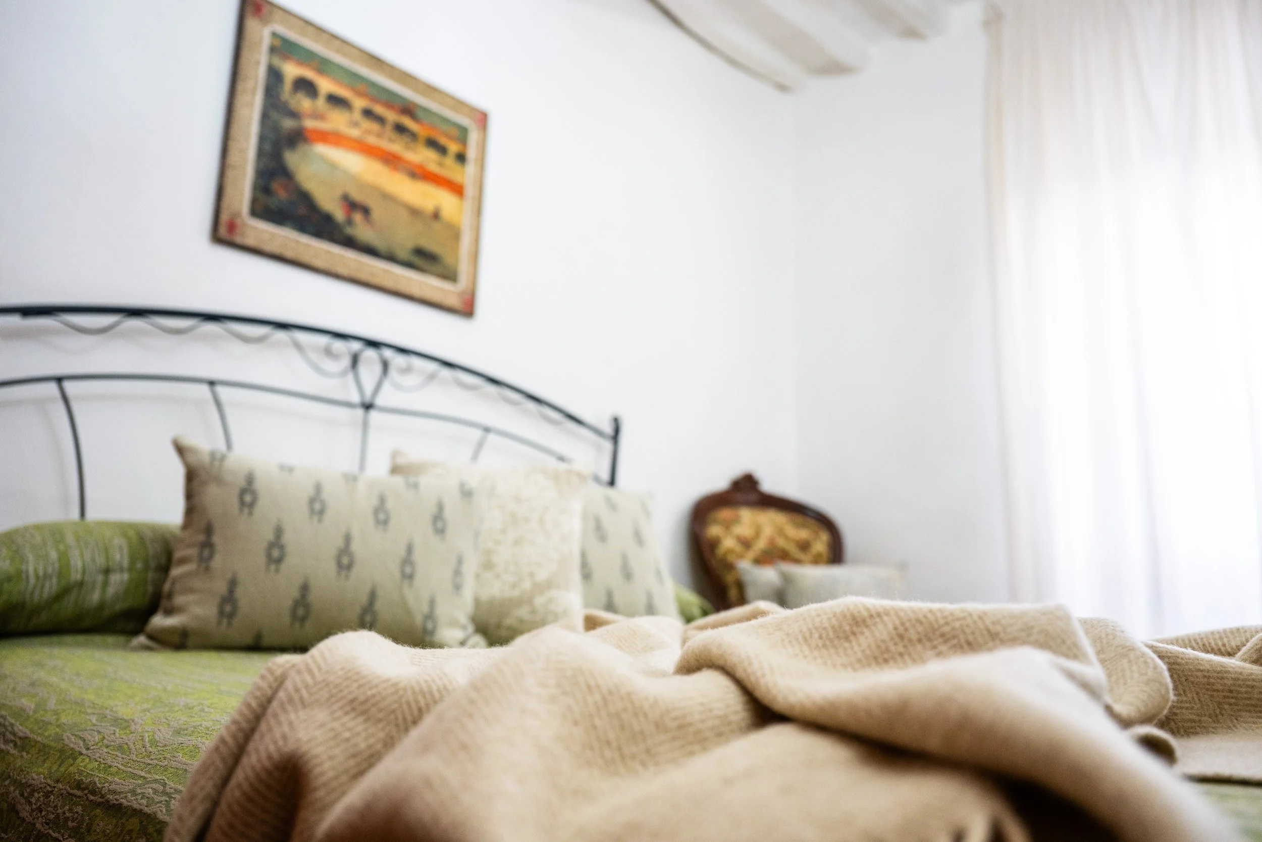 Letto matrimoniale con cuscini e coperta in una stanza luminosa, con una cornice con dipinto sulla parete e una sedia imbottita sullo sfondo