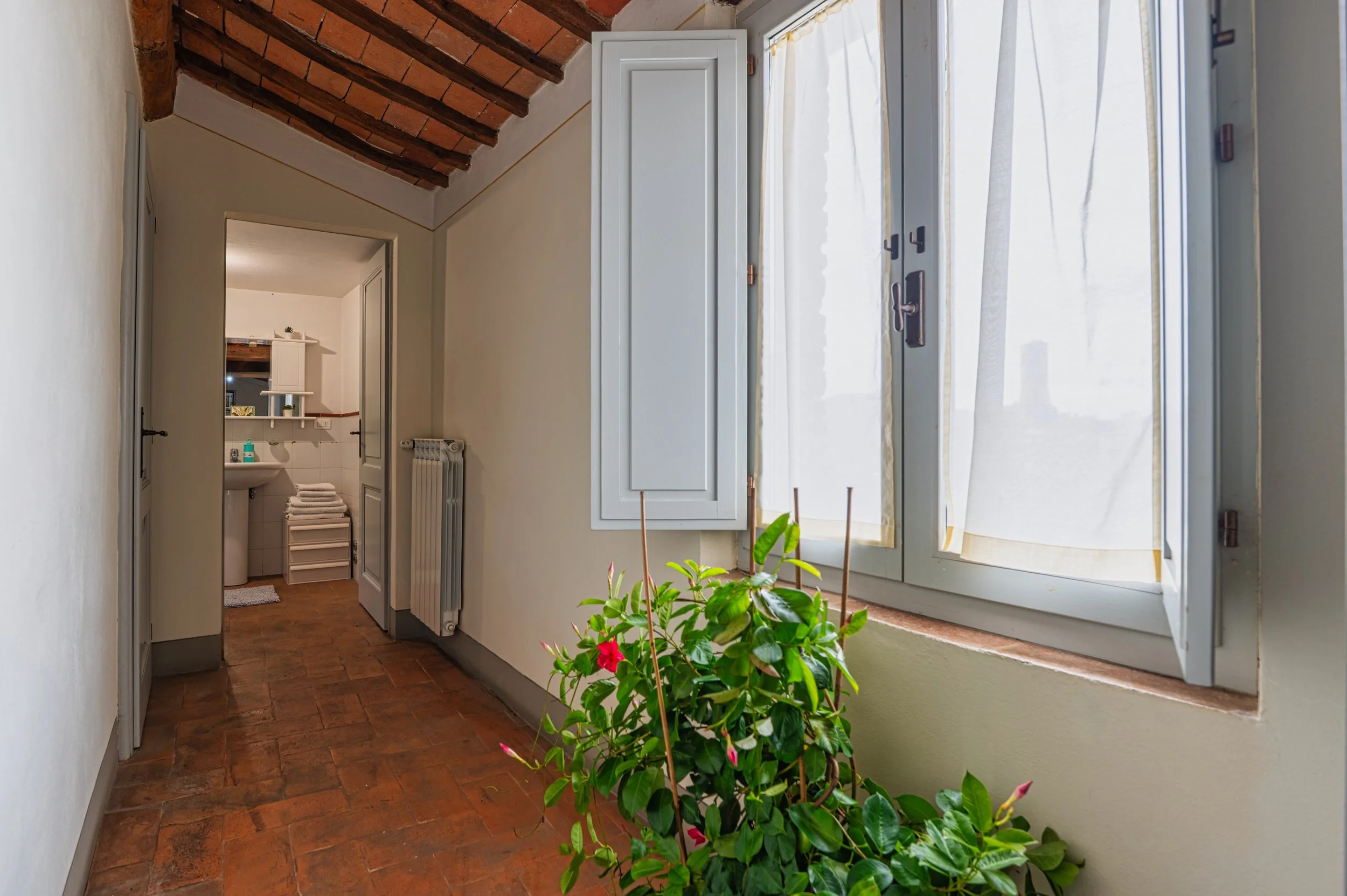 Vista di un corridoio con pareti bianche, pavimento in piastrelle rosse e soffitto in mattoni a vista, con una finestra apribile con tende leggere e un vaso di piante vicino alla finestra, in fondo si vede un bagno con lavabo, specchio e asciugamani.