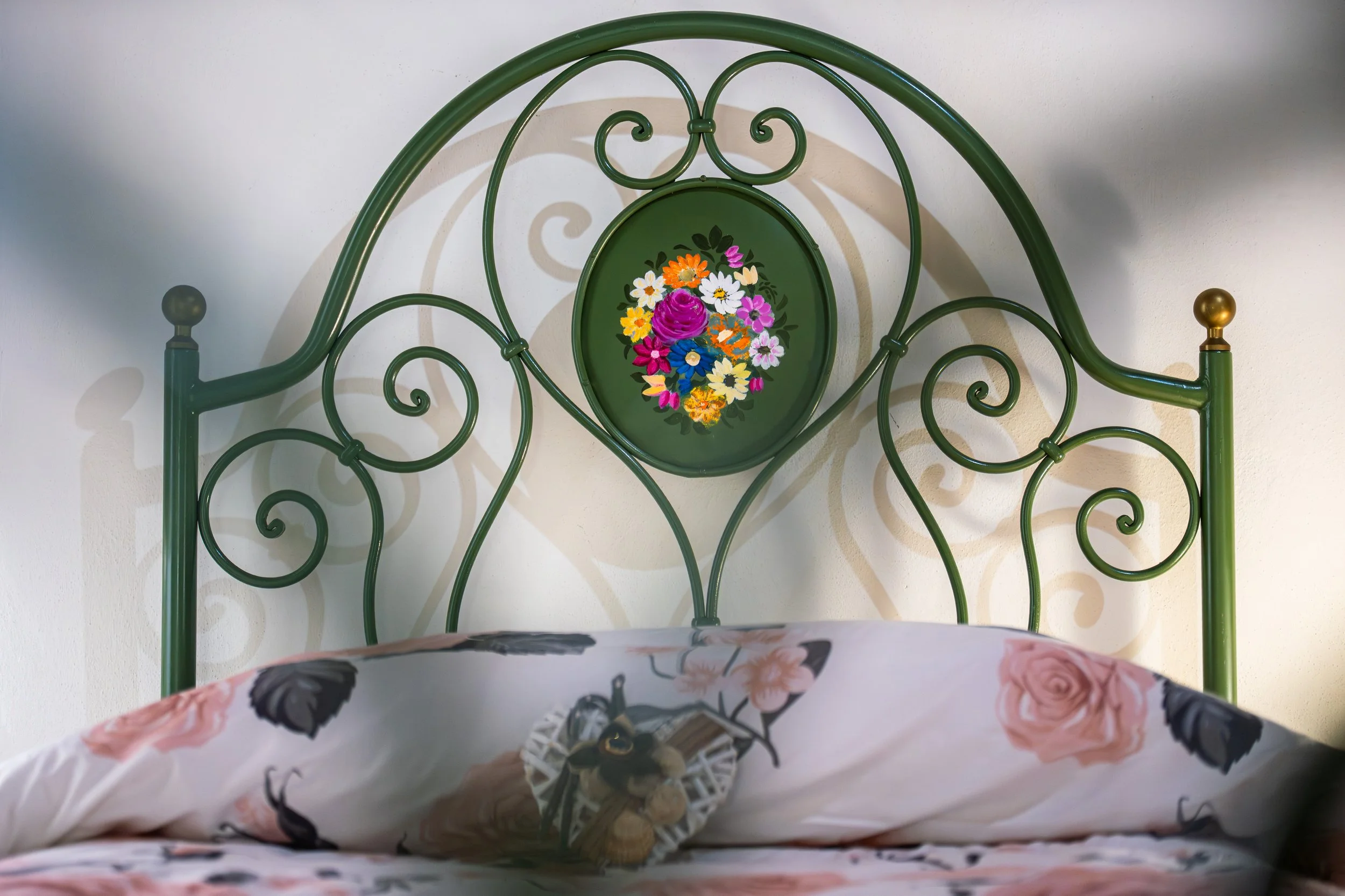 Testiera di ferro verde con motivo a cuori, decorata con un quadro di fiori colorati, situata dietro un letto con copriletto con fiori rosa e grigi.