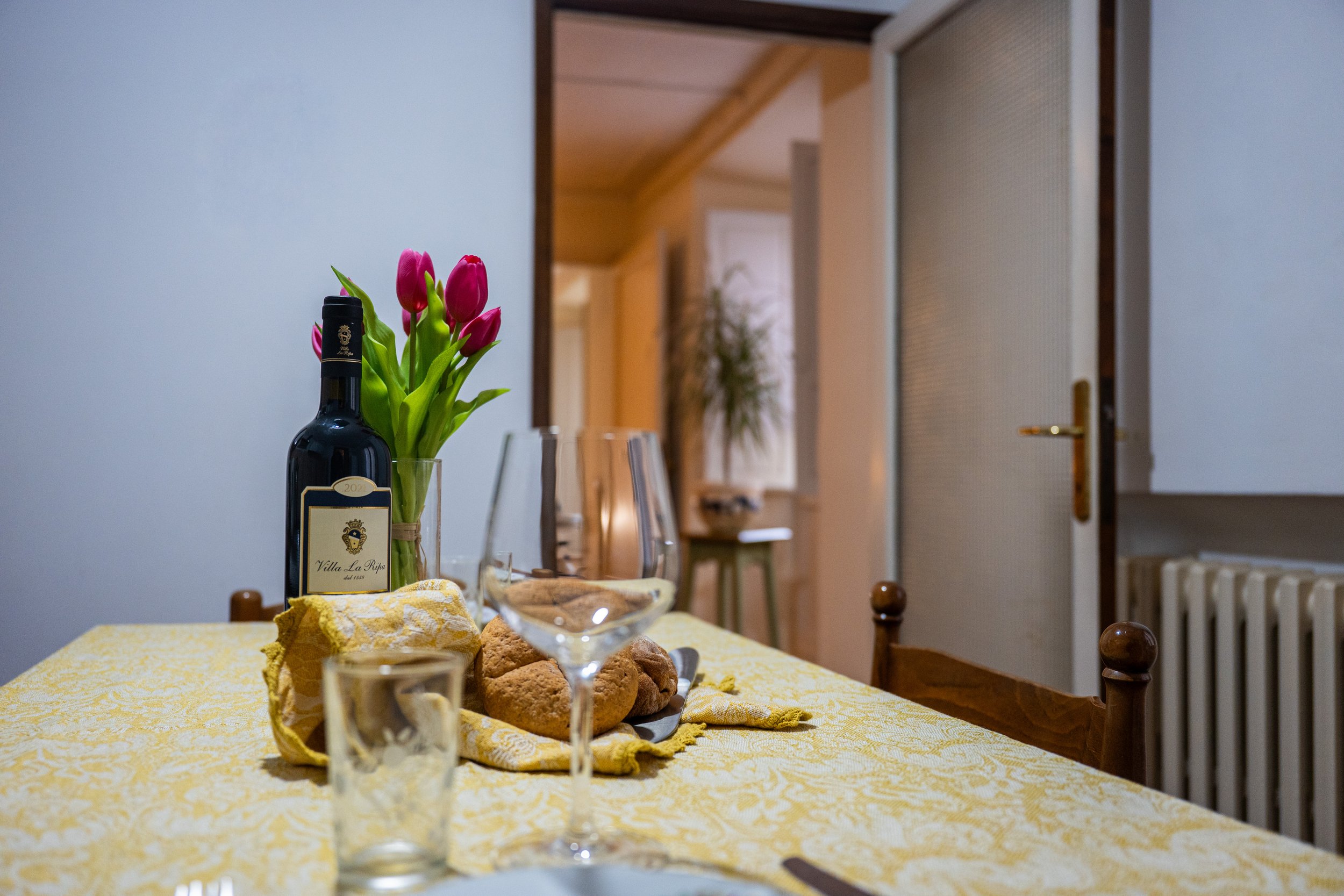 Una tavola apparecchiata con un vaso di tulipani rosa, una bottiglia di vino, alcuni bicchieri, pane e tovaglioli gialli. Sfondo di una stanza con pareti bianche e una porta socchiusa.