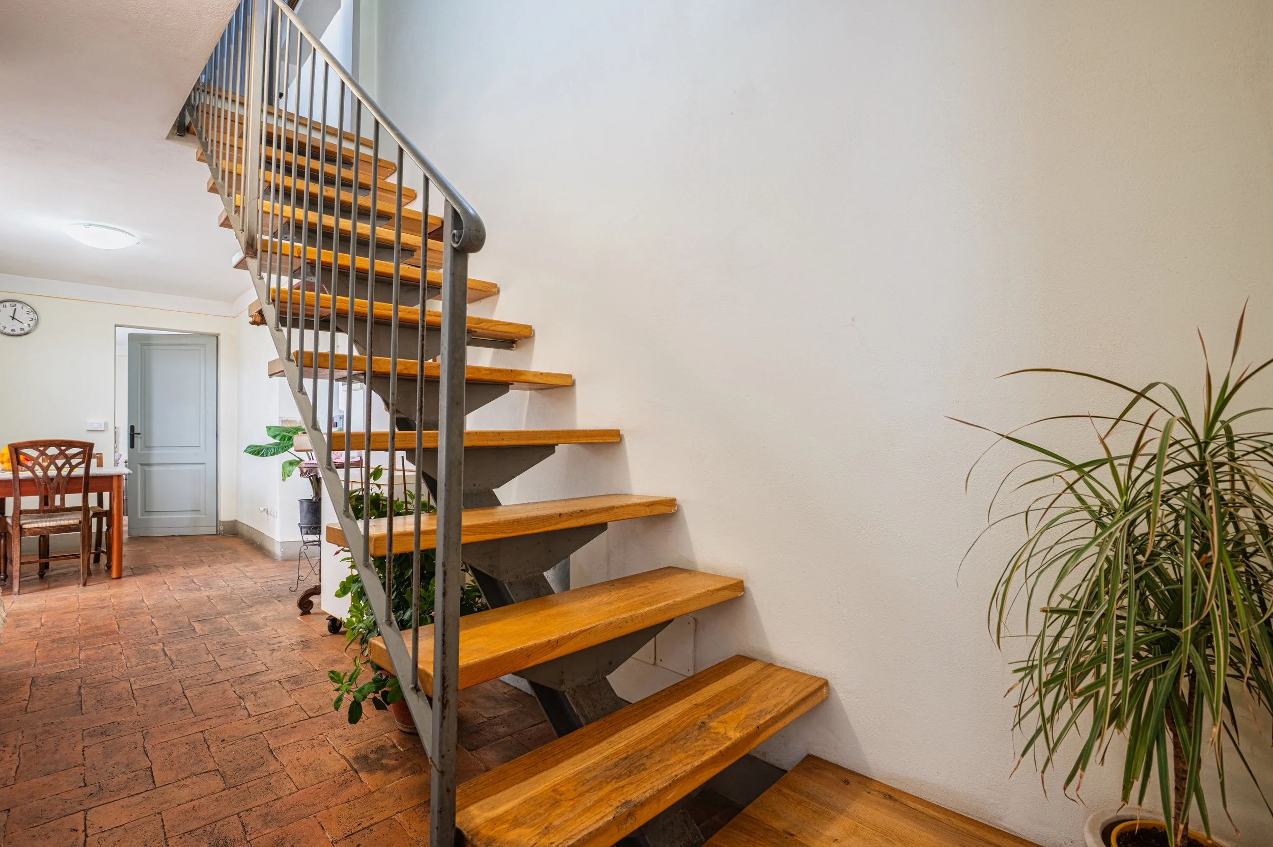 Interno con scala in legno e metallo, pareti chiare, pavimento in piastrelle in terracotta, angolo con piante e tavolo con sedie.