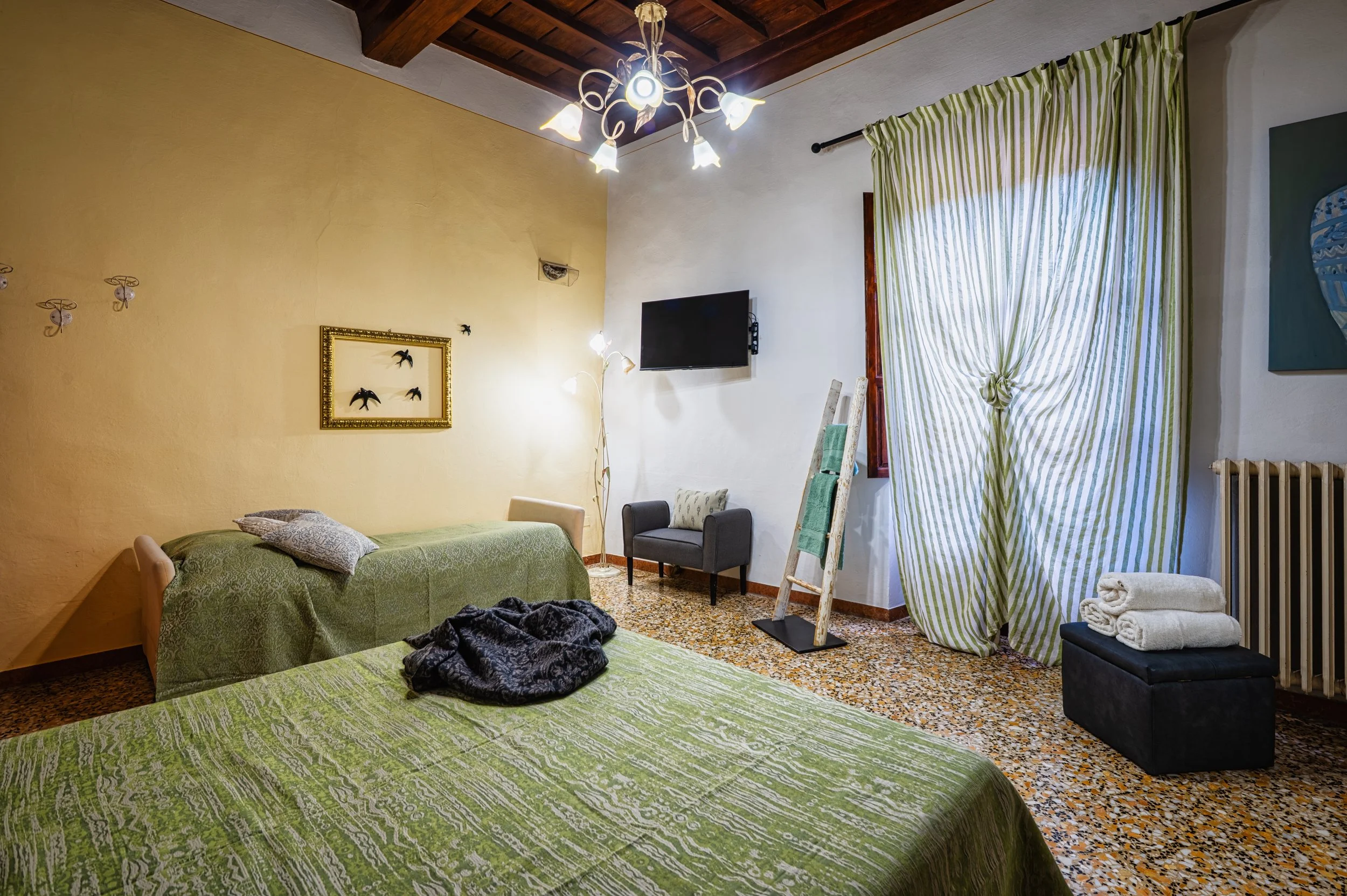 Camera da letto con pareti gialle e bianche, letto con coperta verde, e divano con cuscino. Decorazioni alle pareti, illuminazione a soffitto e lampada da terra. Finestre con tende a righe verdi e bianche, vasca di calorifero e oggetti come asciugamani e quadri.