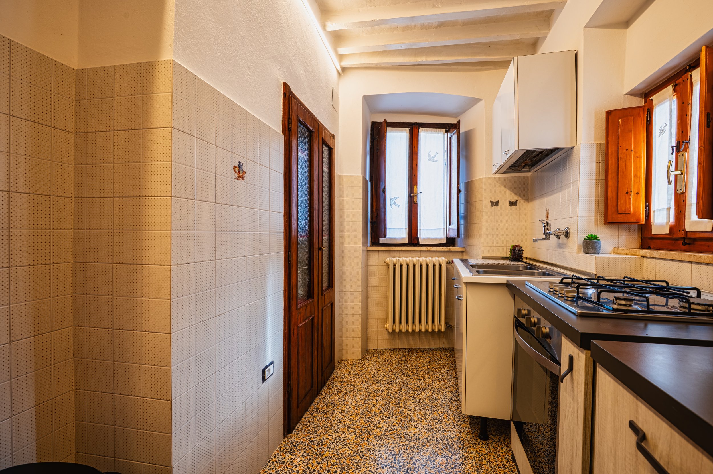 Cucina con pareti rivestite con piastrelle beige, due finestre con tende di cotone e cornici di legno, pensili di legno, piano di lavoro in legno, doppio lavello con rubinetto, gas con fornello e forno, termosifone sotto le finestre, pareti alte soff