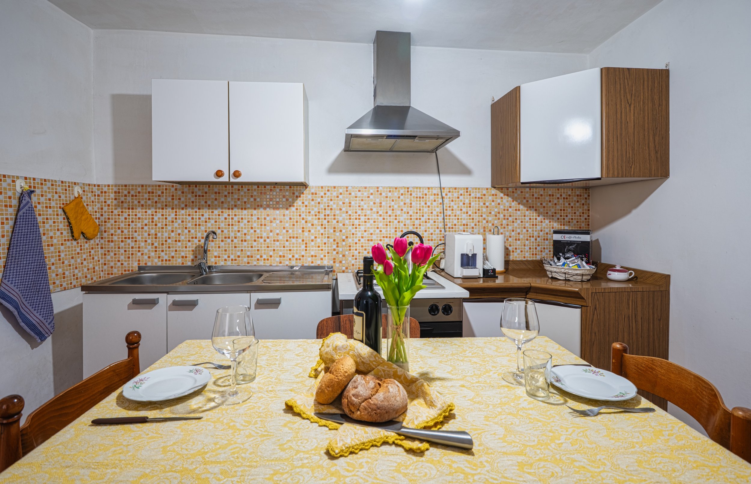Cucina con tavolo apparecchiato con fiori, pane e bottiglia di vino, cucina con lavello, fornelli, armadietti, cappa, e vari elettrodomestici.