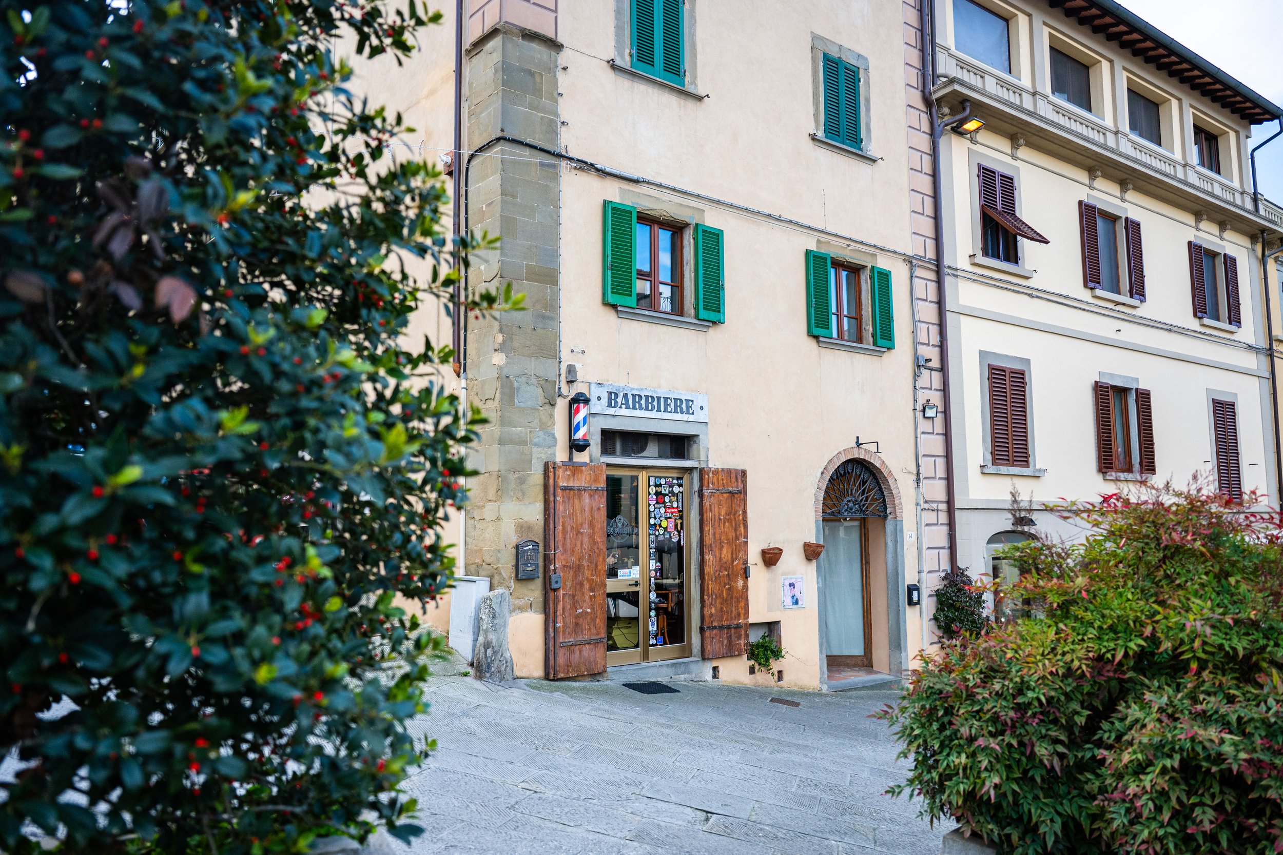 Edificio con pareti color crema e finestre con persiane verdi e marroni, entrata con porte di legno, e piante verdi e arbusti nel cortile