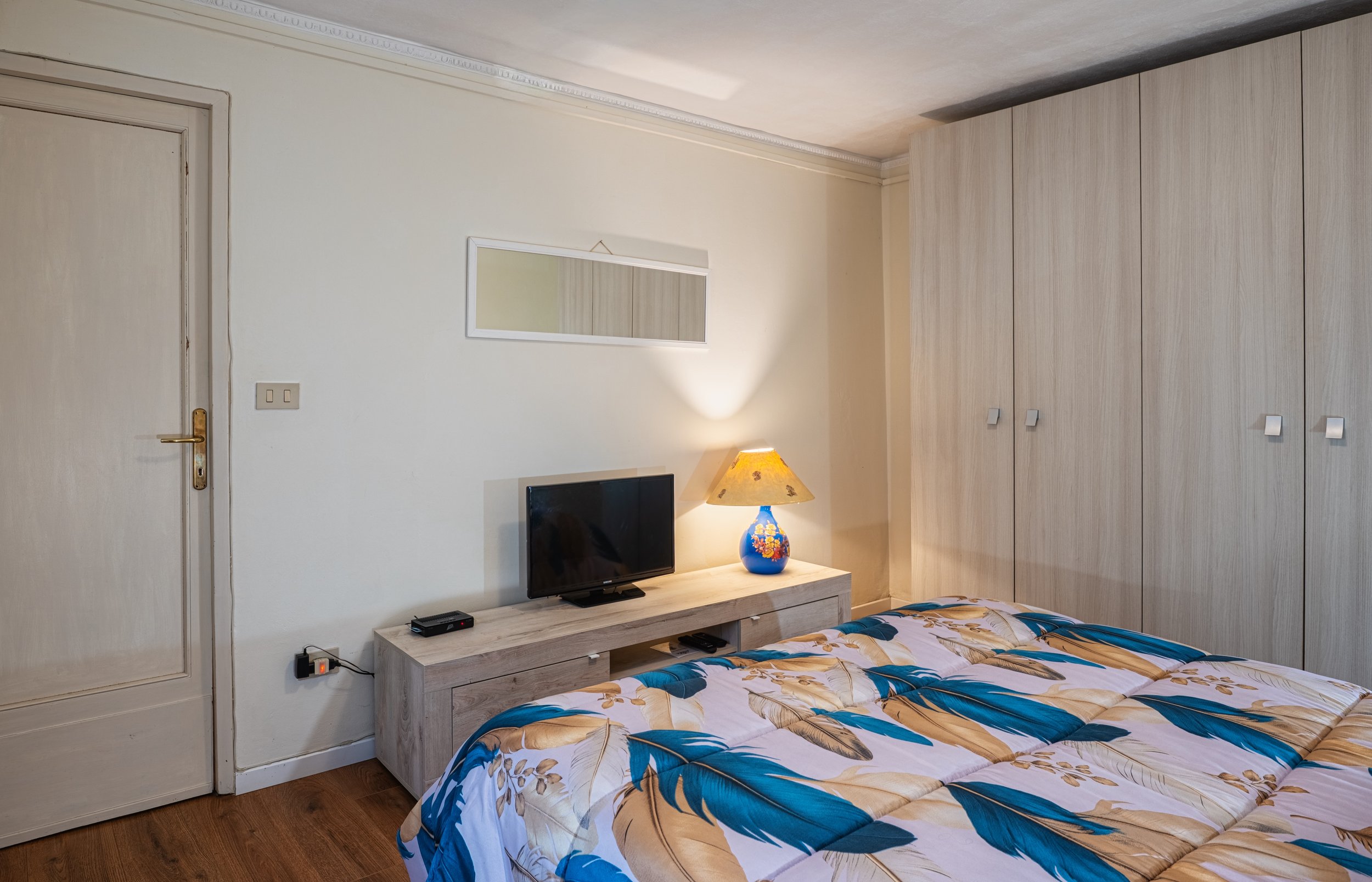 Camera da letto con letto a veiso con coperta colorata, armadio in legno chiaro, piccola televisione sul tavolino, lampada con base blu e paralume con fiori, specchiera rettangolare sulla parete, pareti beige con galleria decorativa e lampada da tavo